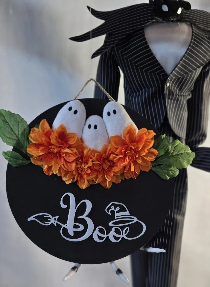 Halloween decor in a black and white striped suit holding a round black sign with the words "Boo" and a witch hat illustration. Attached to the sign are three white ghost decorations with black eyes, surrounded by orange flowers and green leaves.