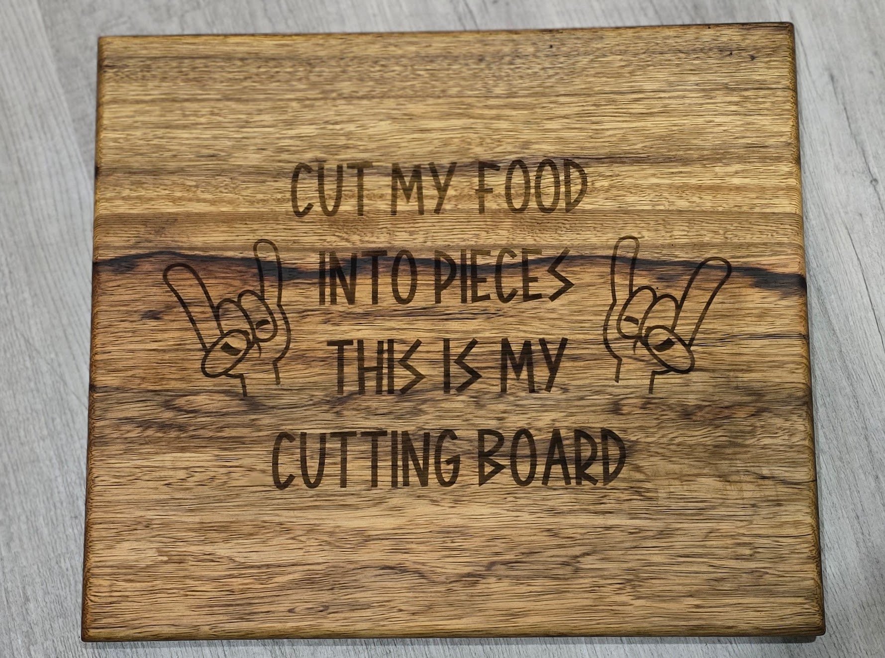 Wooden cutting board with engraved text that reads "Cut my food into pieces this is my cutting board" and illustrations of hands making the "rock on" gesture.