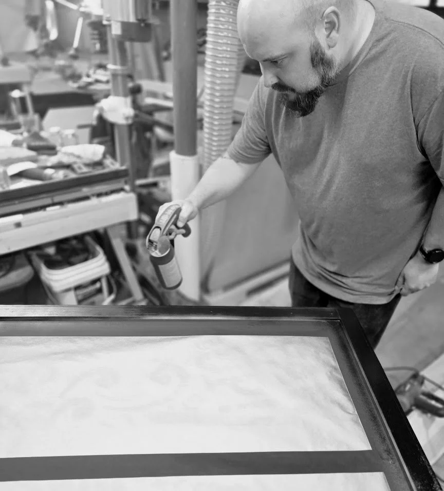 A man in a workshop using a paint spray can on a large, framed piece of wood.