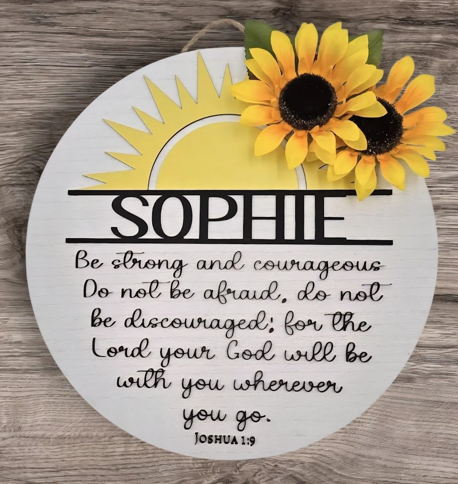 Decorative round wall hanging with a sun illustration and the word 'SOPHIE' in bold black letters, featuring a Bible verse from Joshua 1:9 written in cursive, adorned with a bunch of three yellow sunflowers in the top right corner.
