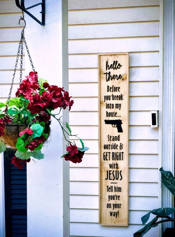 A vertical wooden sign with black text and a gun silhouette, mounted on white house siding. The sign says: 'hello there. Before you break into my house... Stand outside & GET RIGHT with JESUS Tell him you're on your way!'
