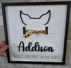 Decorative framed wall art featuring a minimalist outline of a dog's face with pointed ears, a bowtie, and the name 'Addison' below. Text reads 'You Left Paw Prints on Our Hearts'.