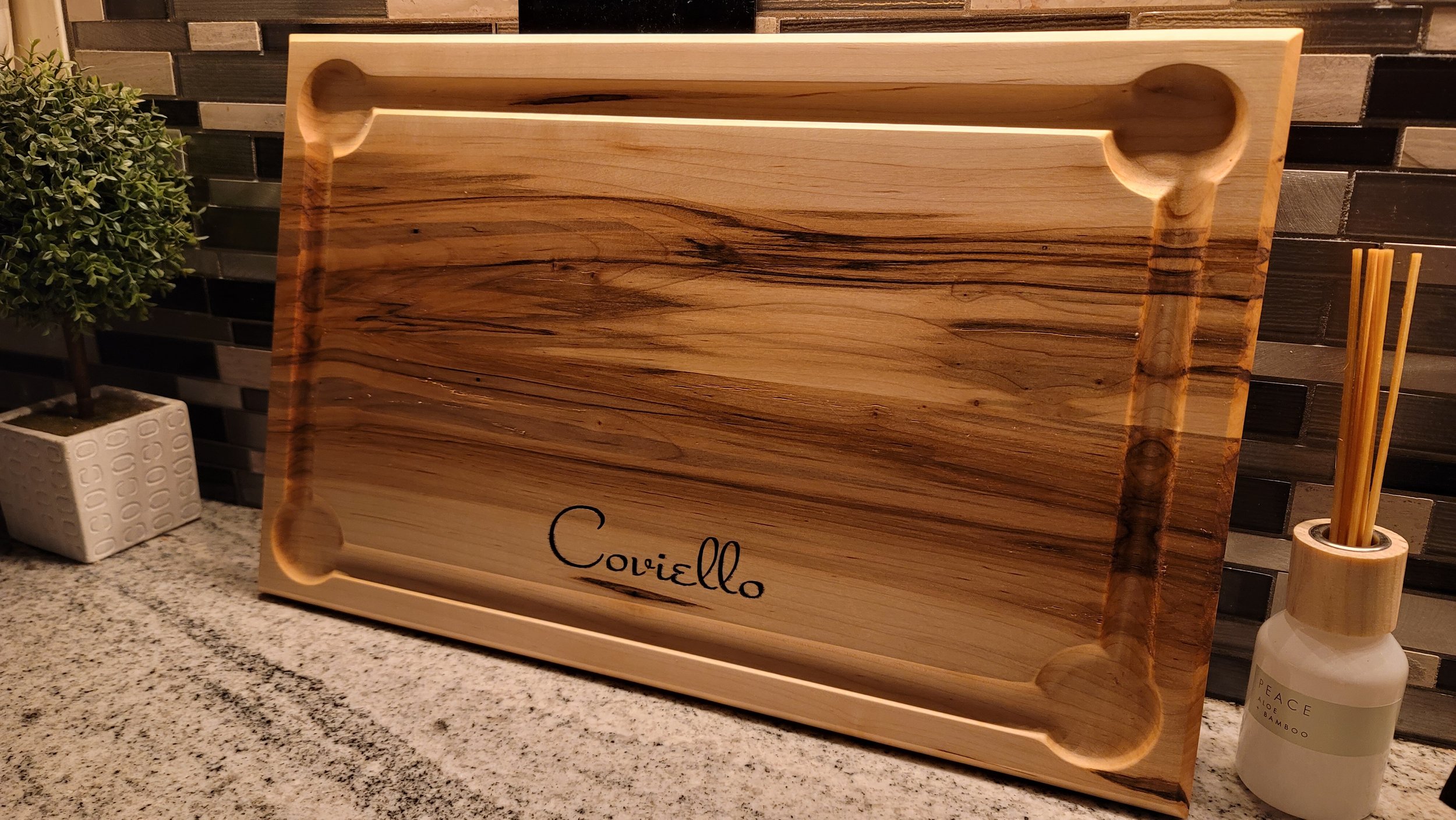 Wooden serving tray with the name 'Coviello' engraved on it, placed on a speckled granite countertop, with a potted plant and a bottle of reed diffusers nearby.