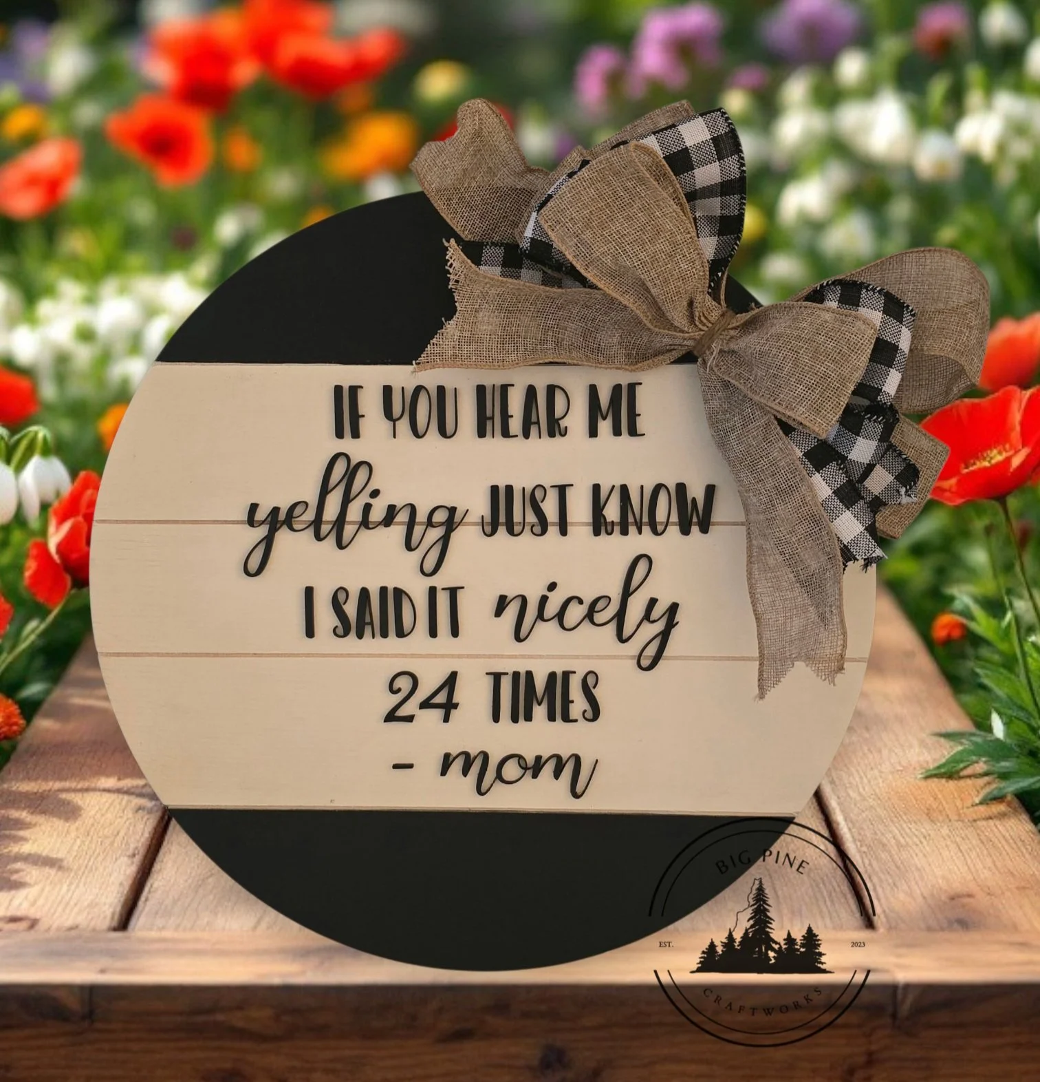 Wooden sign with humorous message about talking loudly, decorated with a burlap and checkered ribbon bow, placed on a wooden surface with colorful flowers in the background.