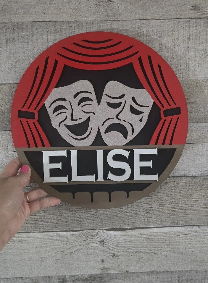 A decorative plaque with theatrical masks, one laughing and one crying, beneath a red curtain, and the name Elise at the bottom.