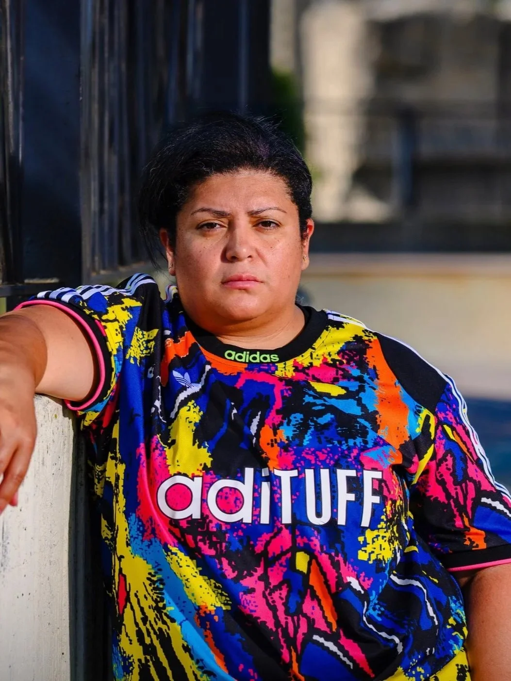 A woman wearing a colorful Adidas sports shirt taking a selfie outdoors with a serious expression.