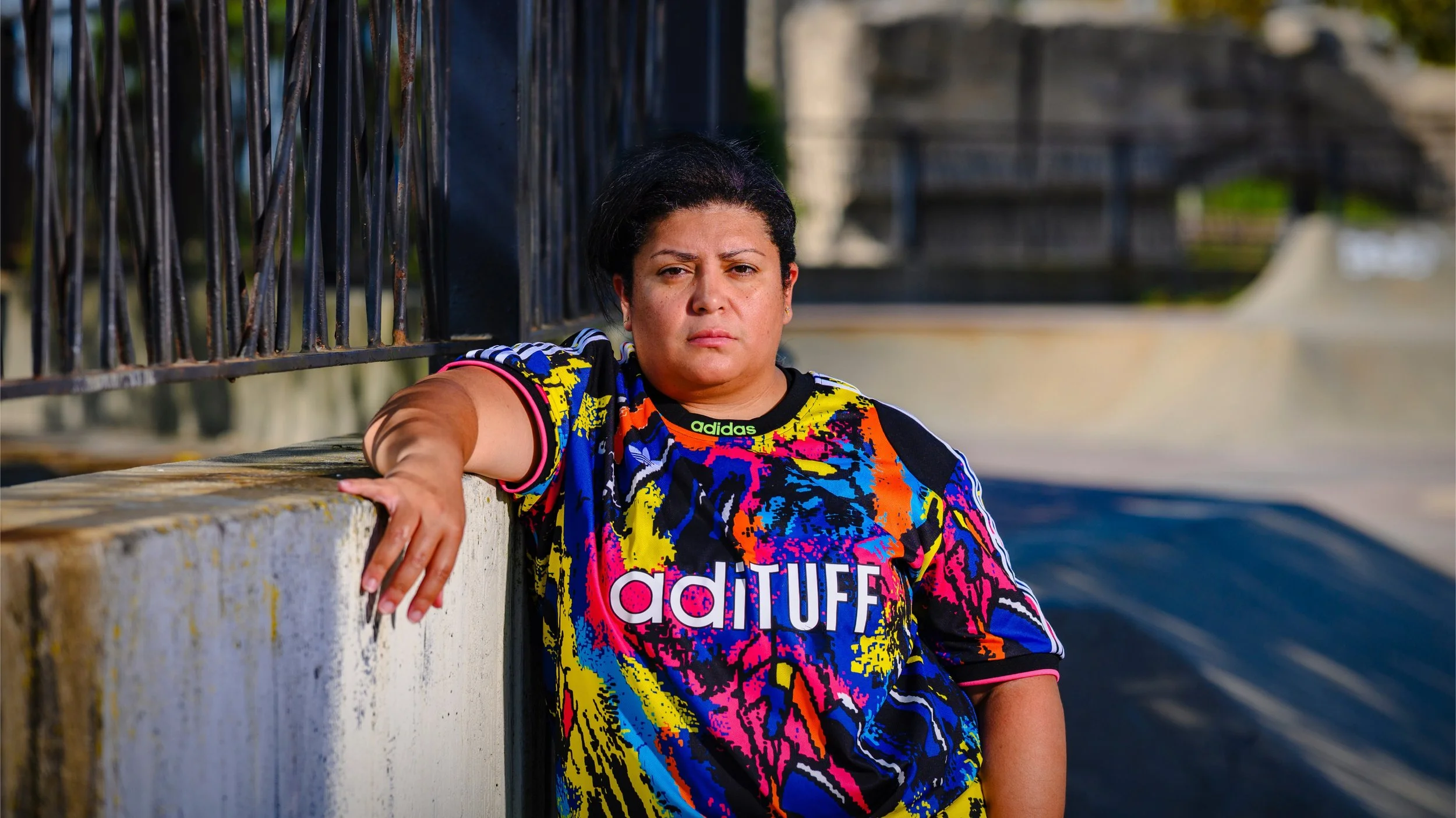 A woman with dark hair and medium skin tone leaning on a concrete barrier, wearing a colorful Adidas sports shirt with the word 'aditUFF' and a black background, outdoors near a skate park or similar structure, with a serious expression on her face.