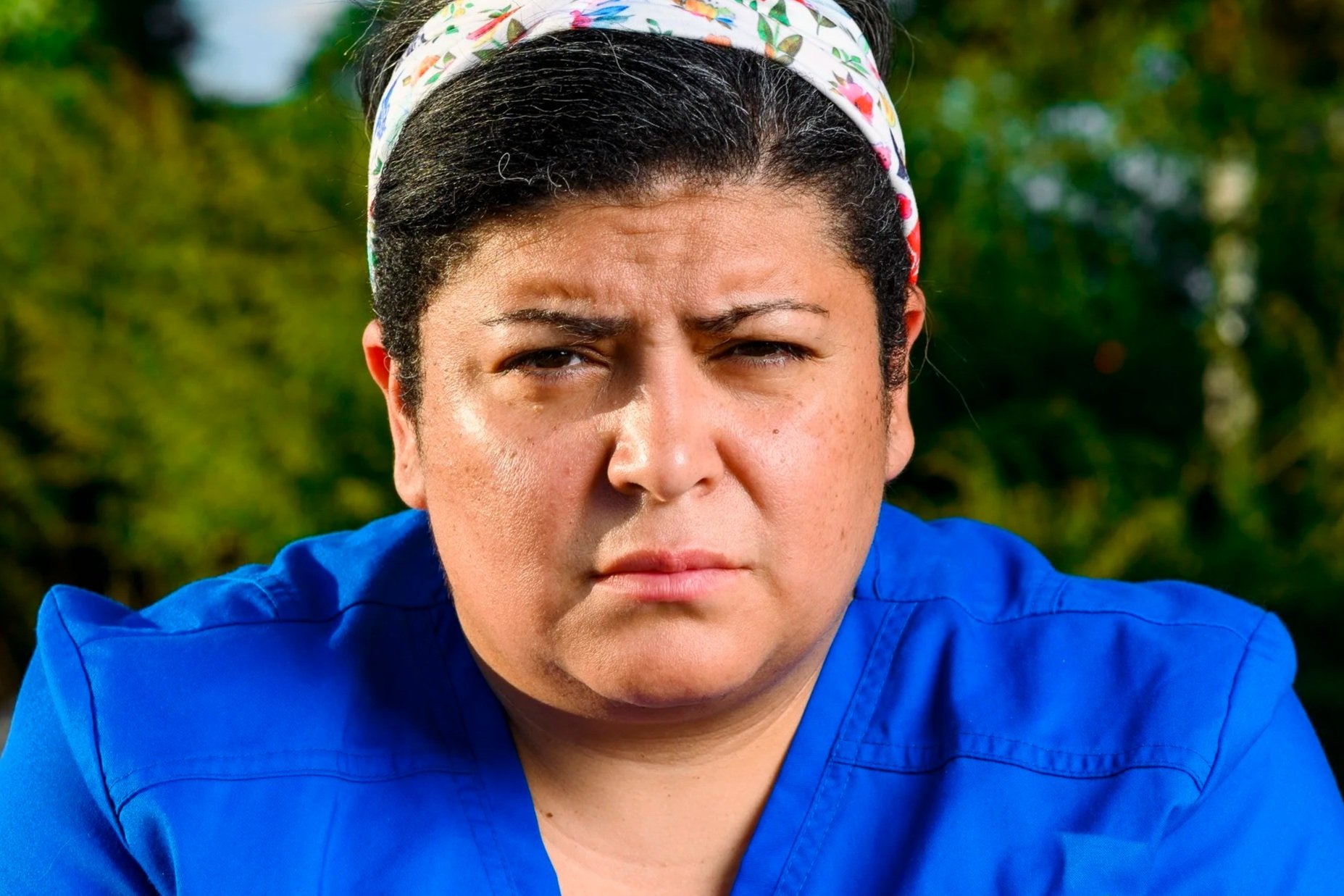 A woman with short dark hair wearing a floral headband and blue scrubs, outside with blurred green foliage in the background.