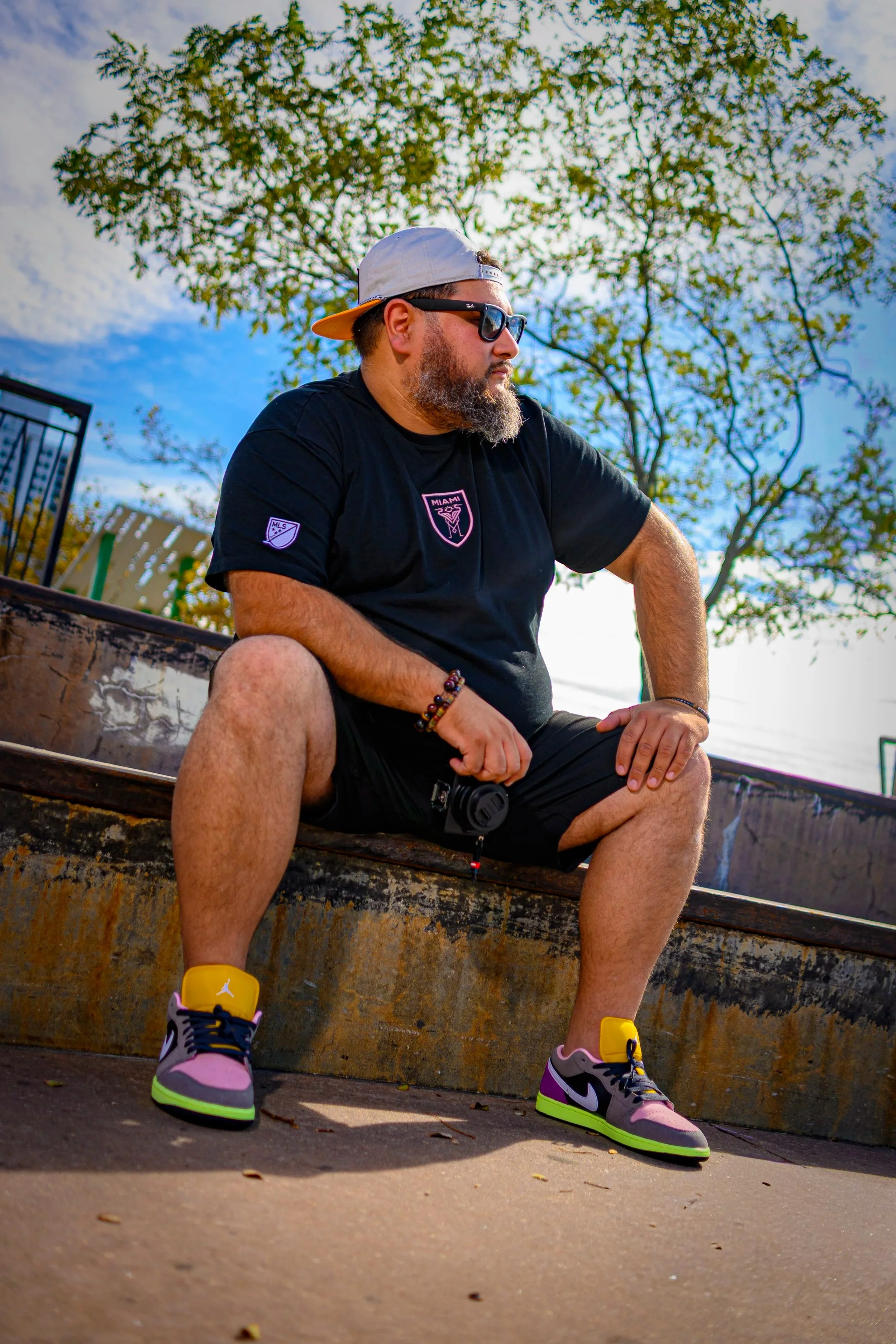 A bearded man wearing glasses, a baseball cap, and a white T-shirt with a logo, standing outdoors with trees and city buildings in the background.
