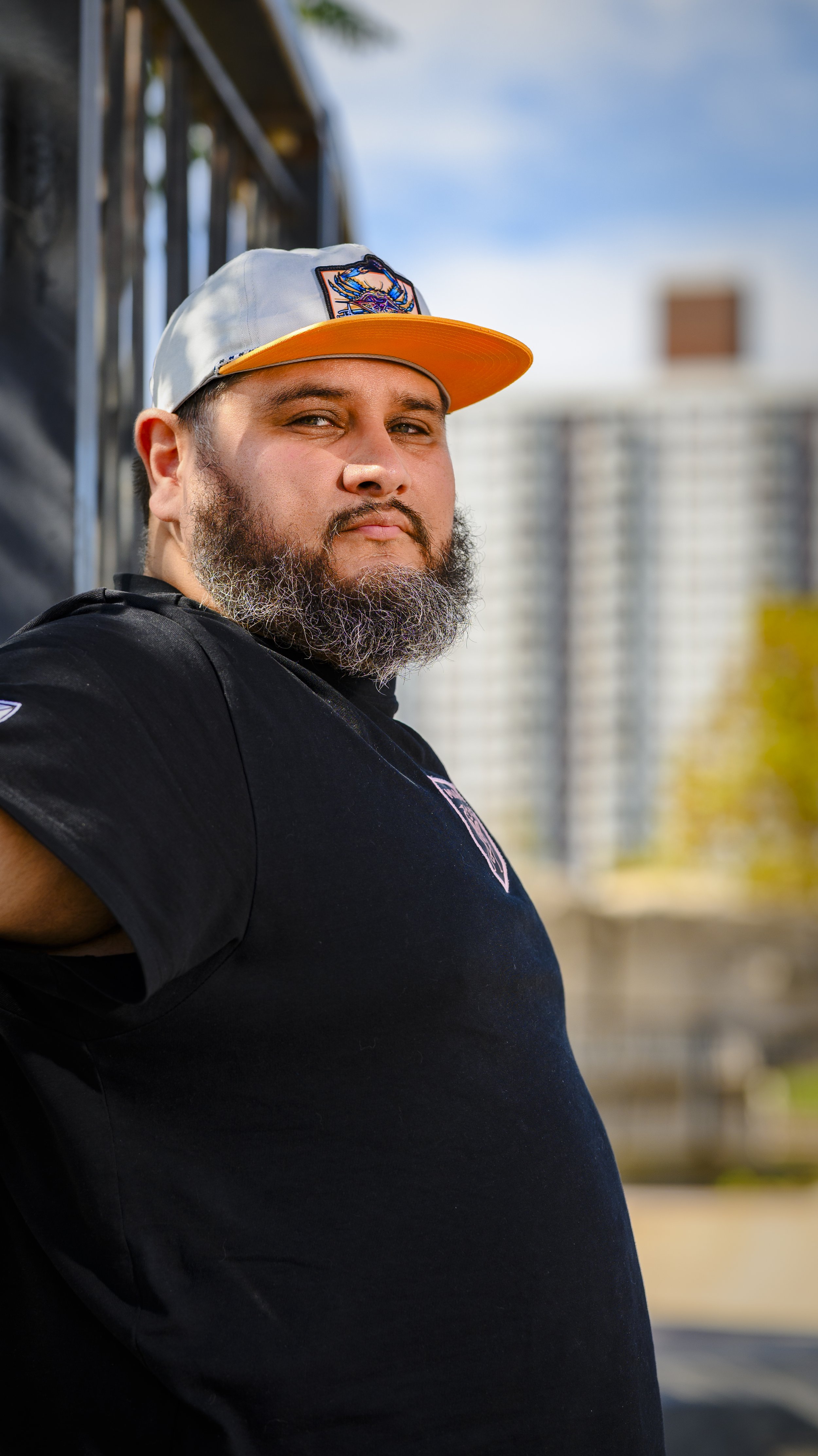 A man with a beard wearing a black t-shirt and a gray baseball cap with an orange brim, standing outdoors with blurred buildings and sky in the background.