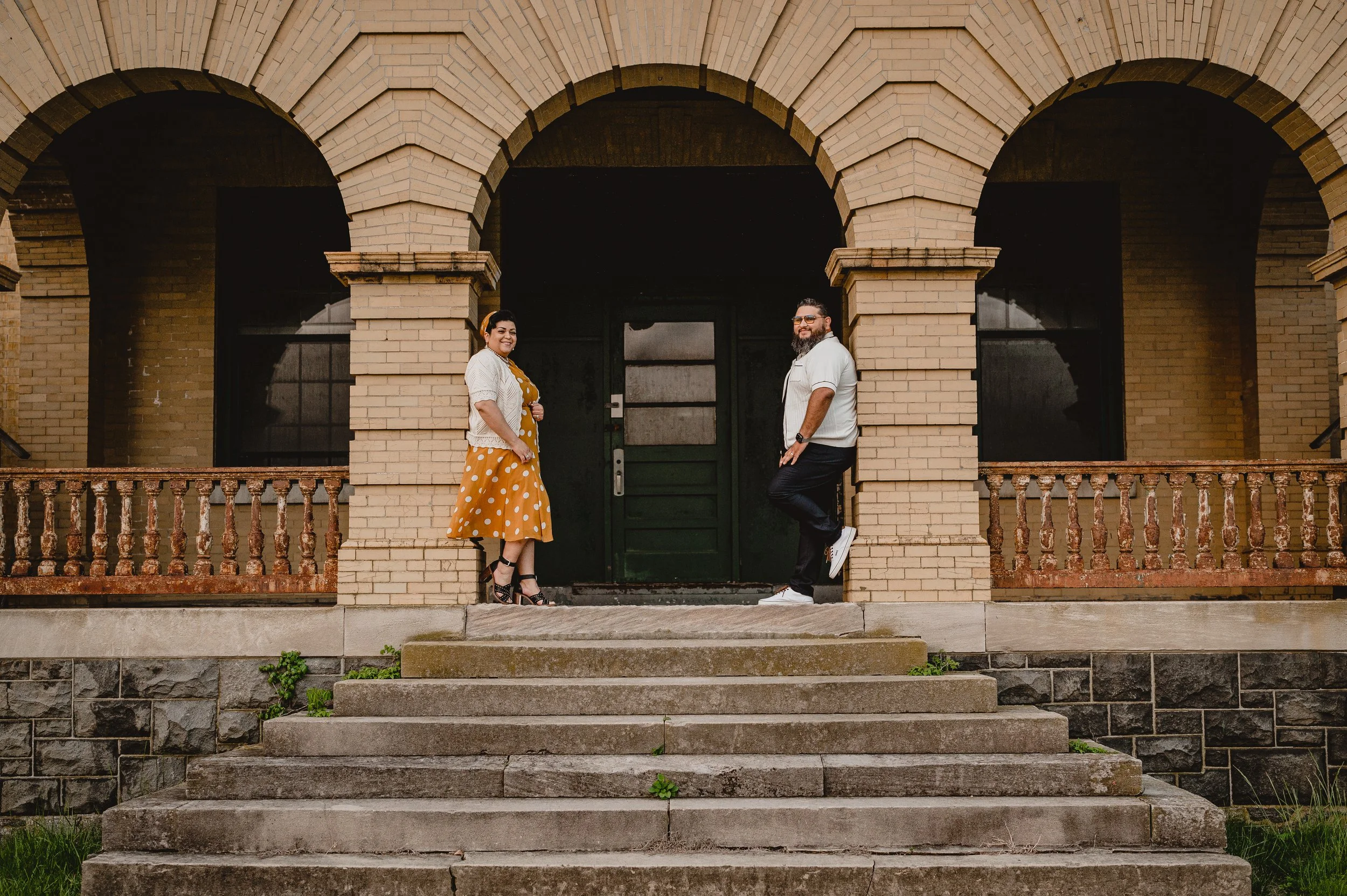 A woman in a mustard yellow polka dot dress and a white cardigan stands on the left, and a man in a white shirt and dark pants leans against a column on the right, both smiling in front of an old brick building with archways.
