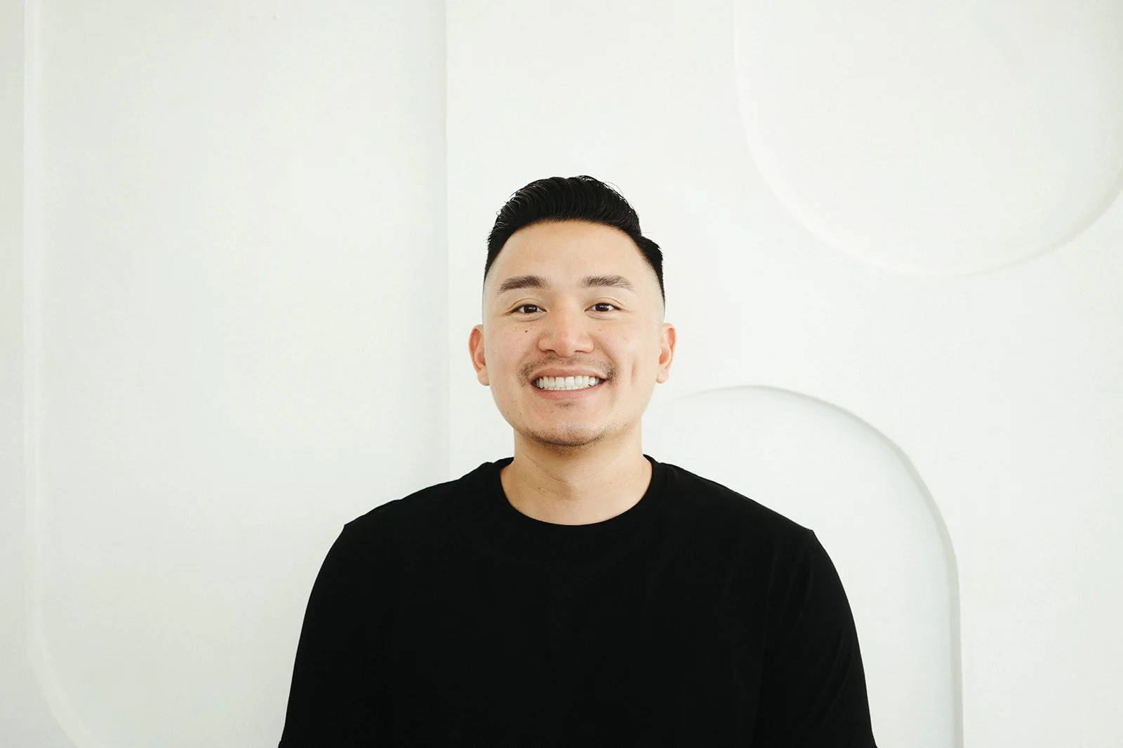 A smiling Asian man with black hair, wearing a black shirt, standing in front of a white wall with circular cut-out designs.