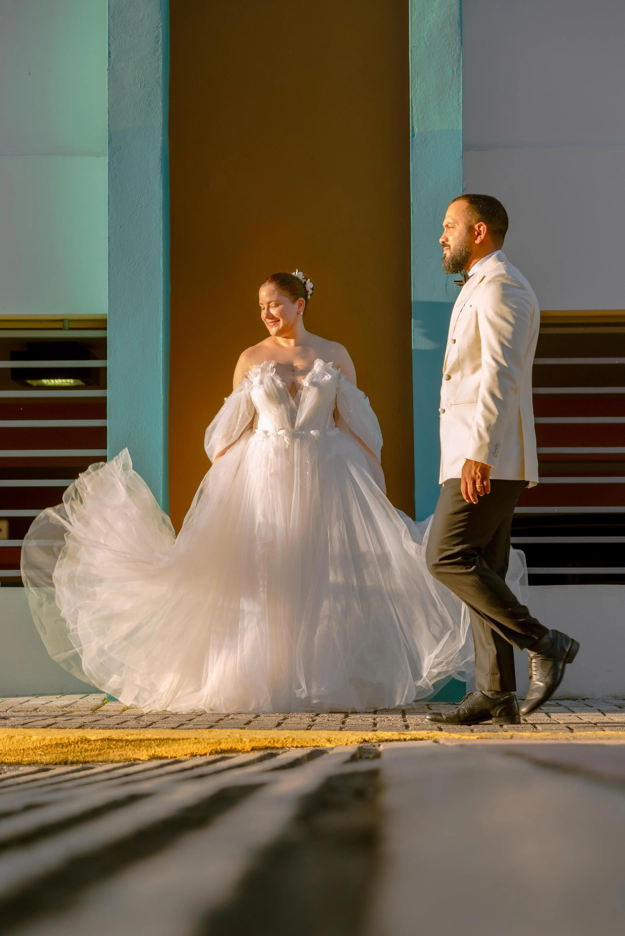 A bride in a white wedding gown smiling and looking down, standing next to a groom in a white tuxedo jacket and black pants, outside a building during sunset in Puerto Rico.