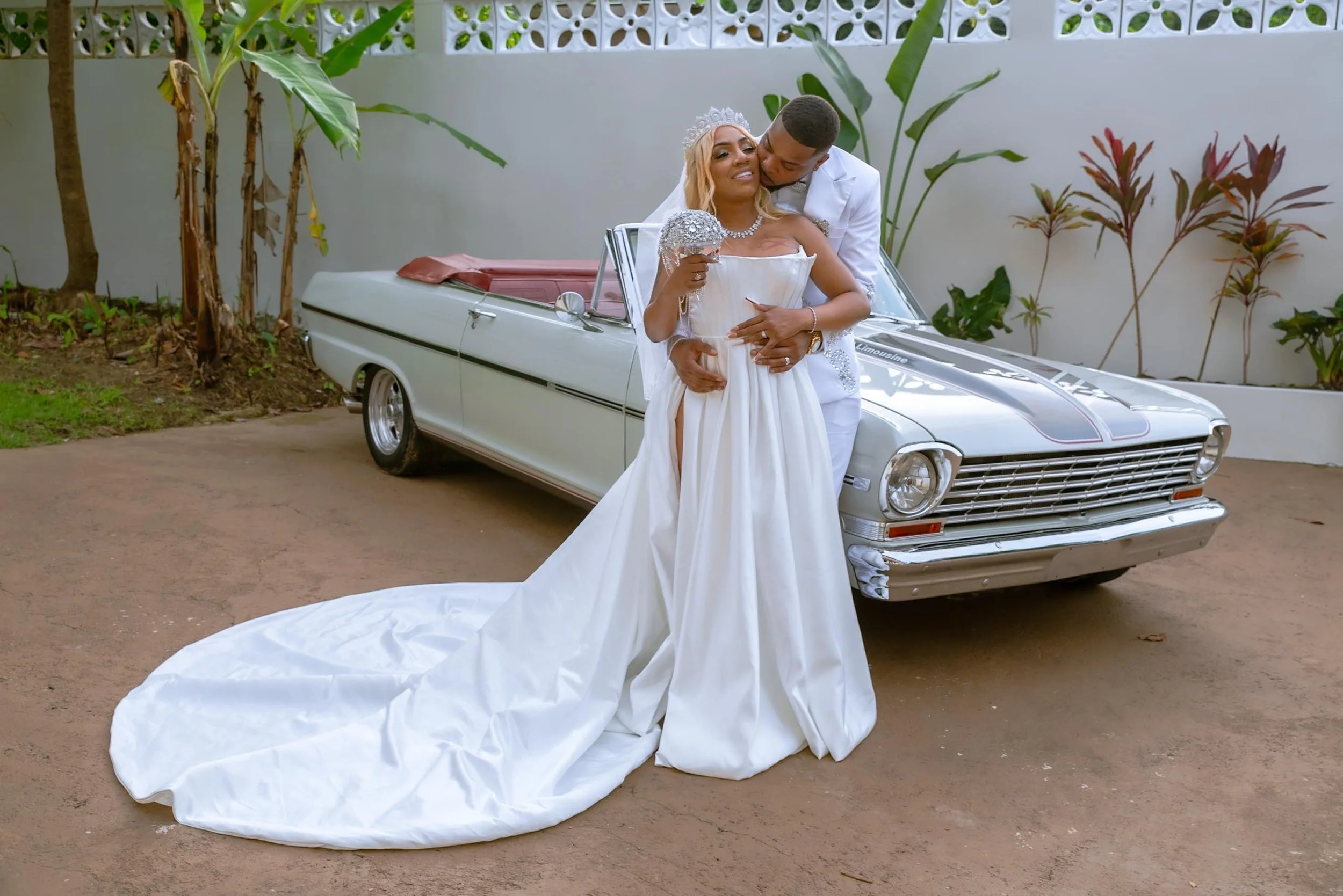 A bride and groom in wedding attire are embracing and kissing in front of a classic white car with a black stripe, parked on a dirt driveway, with tropical plants and a white wall in the background in Puerto Rico.