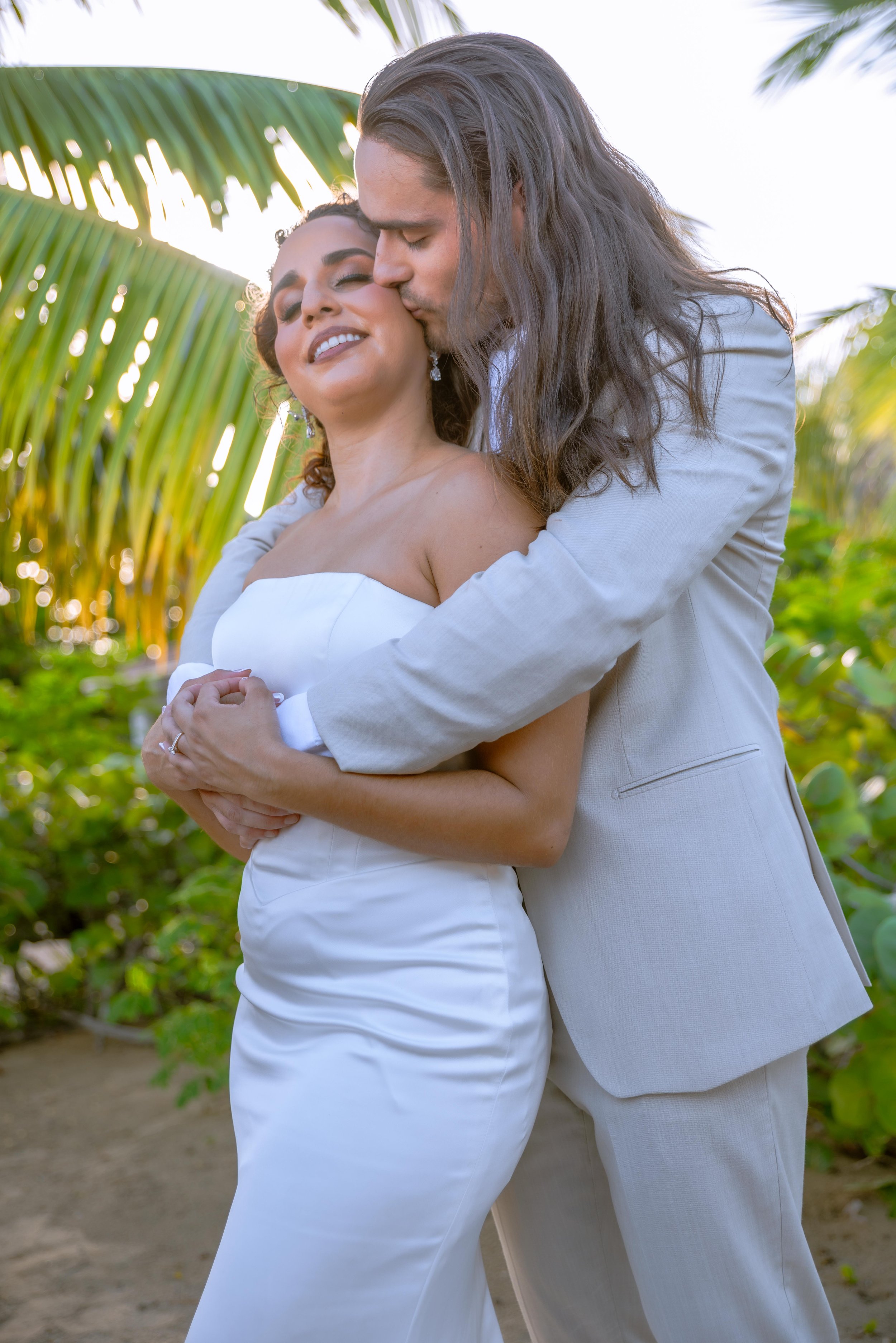 A man in a light-colored suit kisses a woman in a white dress on a tropical beach, with palm trees and greenery in the background in Puerto Rico.