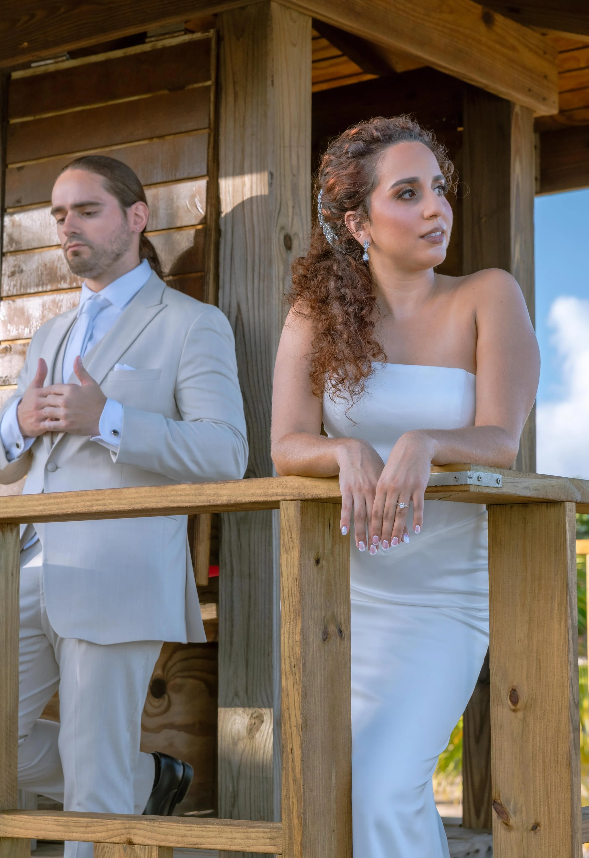 A woman in a white strapless dress with curly hair and earrings leans on a wooden railing, looking into the distance. A man in a light-colored suit with long hair tied back stands behind her with his hands on his chest, looking downward in Puerto Ri.