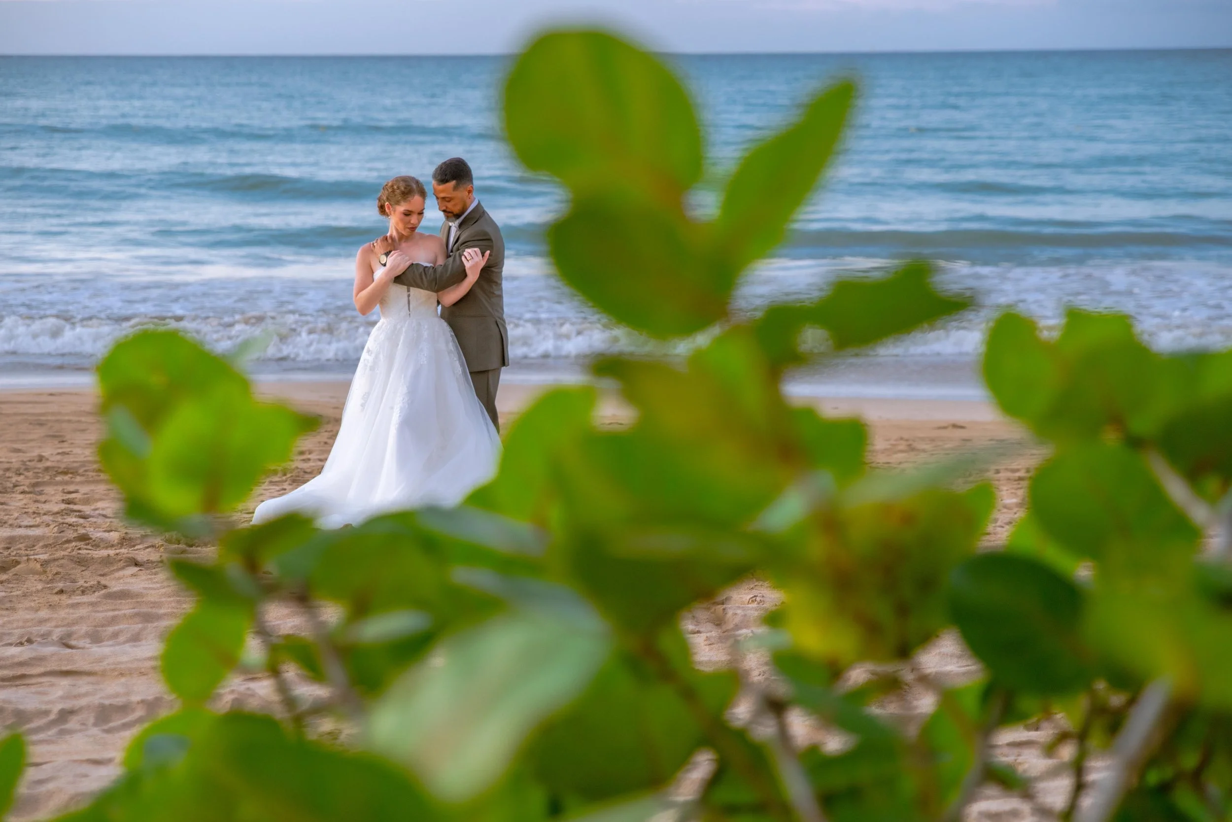 A bride and groom standing on the beach, embracing each other, with the ocean in the background and green foliage in the foreground in Puerto Rico.