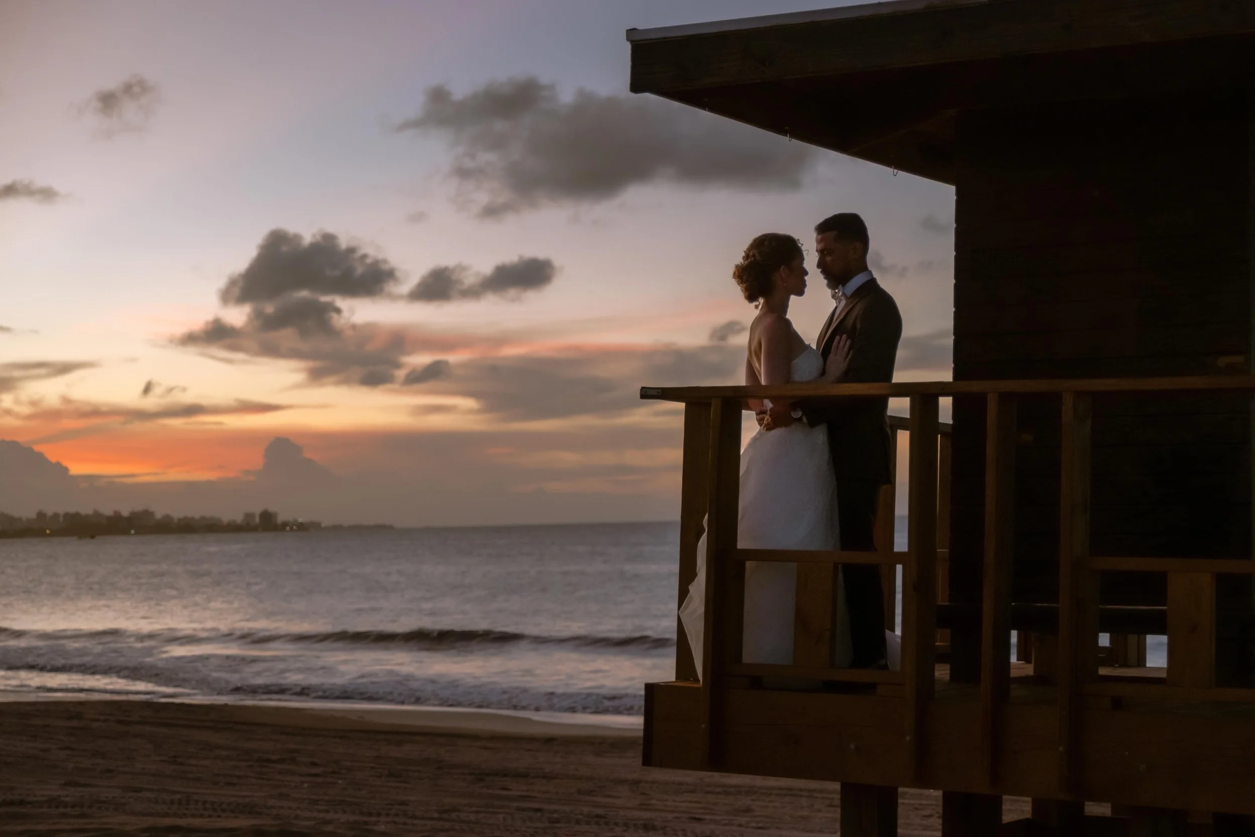 A bride and groom standing close together on a wooden balcony overlooking the beach at sunset, embracing and gazing into each other's eyes in Puerto Rico.