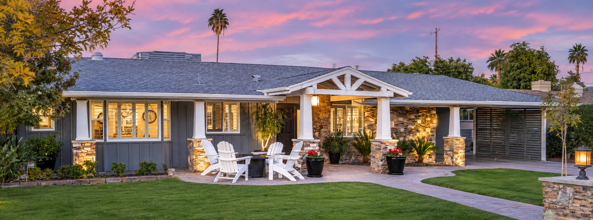 A house with a stone and wood exterior, lit up at dusk with a pink and purple sky in the background. The front yard has a lush green lawn and potted plants near the entrance with white Adirondack chairs around a table on the patio.