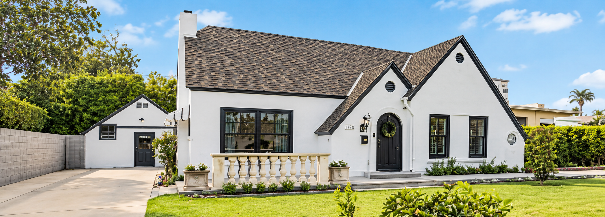White house with black trim, front porch with white balustrade, and a shingled roof, surrounded by a well-maintained lawn and trees, under a blue sky.