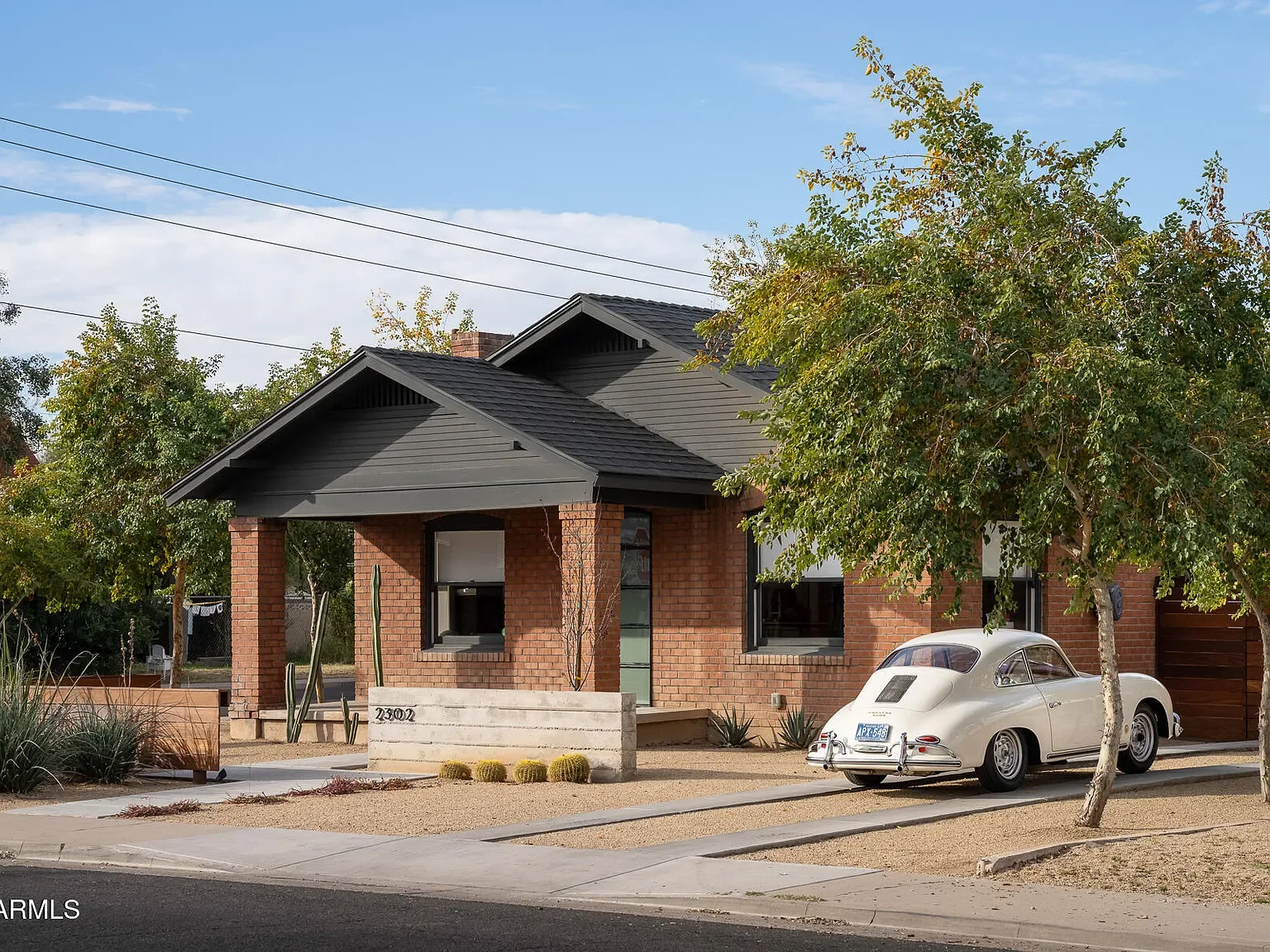 A modern suburban house with a brick exterior, black roof, and large windows. There is a vintage white car parked in the driveway, a tree with green leaves in front, and desert landscaping with cacti and small bushes.