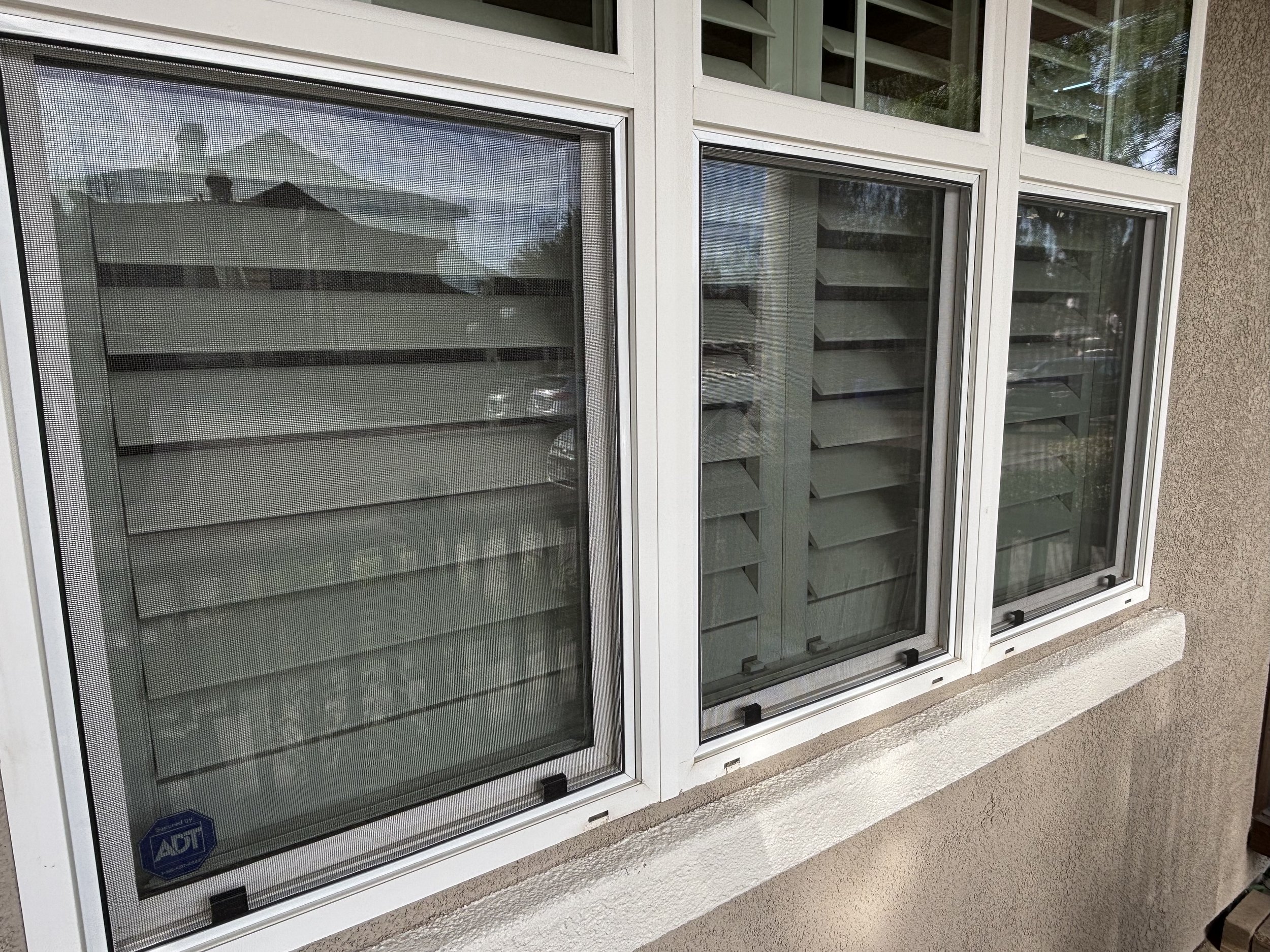 Three-panel casement windows with metal screens and horizontal blinds in a residential building, reflecting nearby structures and trees.