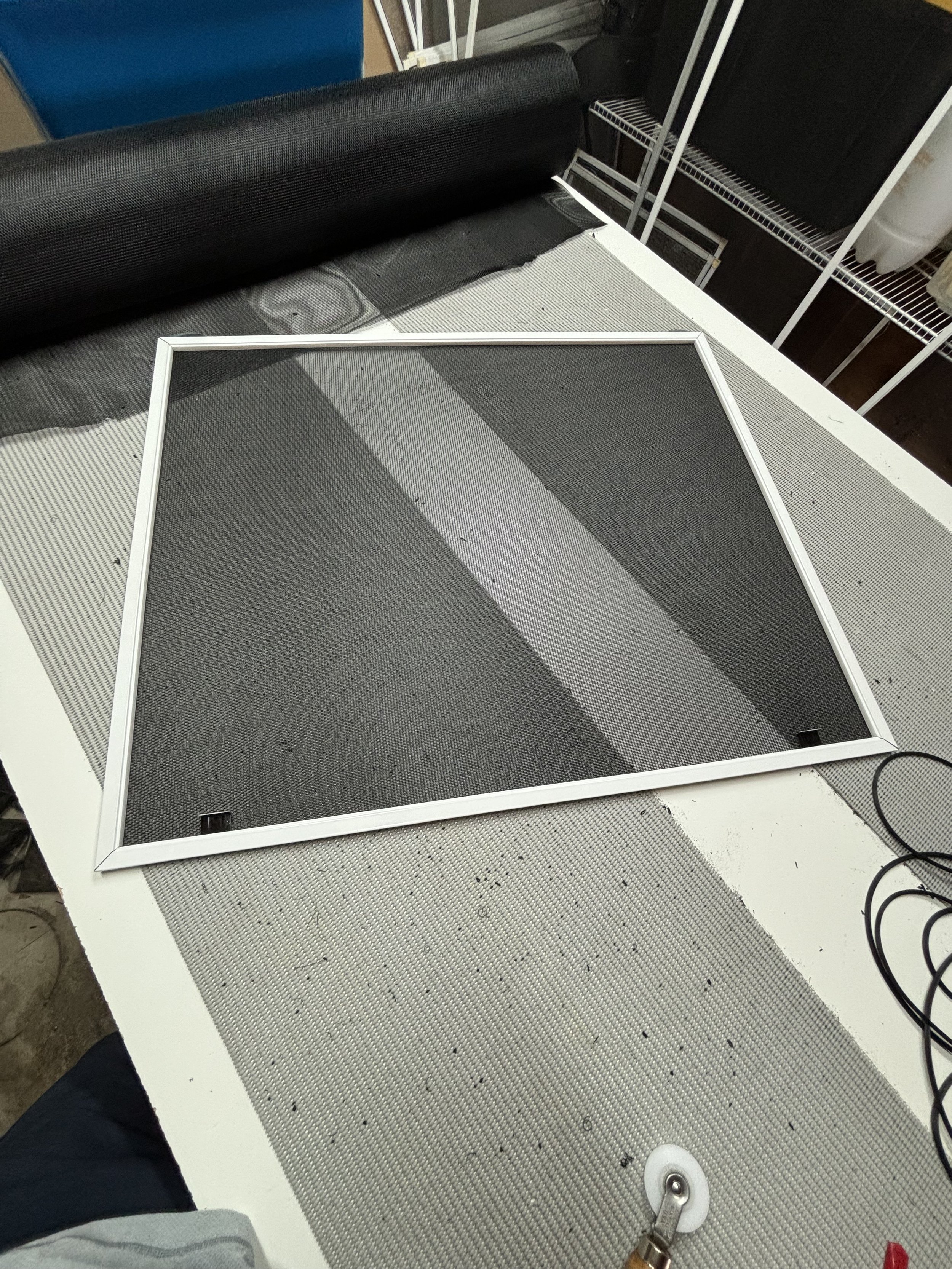 A window screen on a workbench with black mesh and white frame, surrounded by screen repair tools and materials, including a roll of mesh and a spline roller.