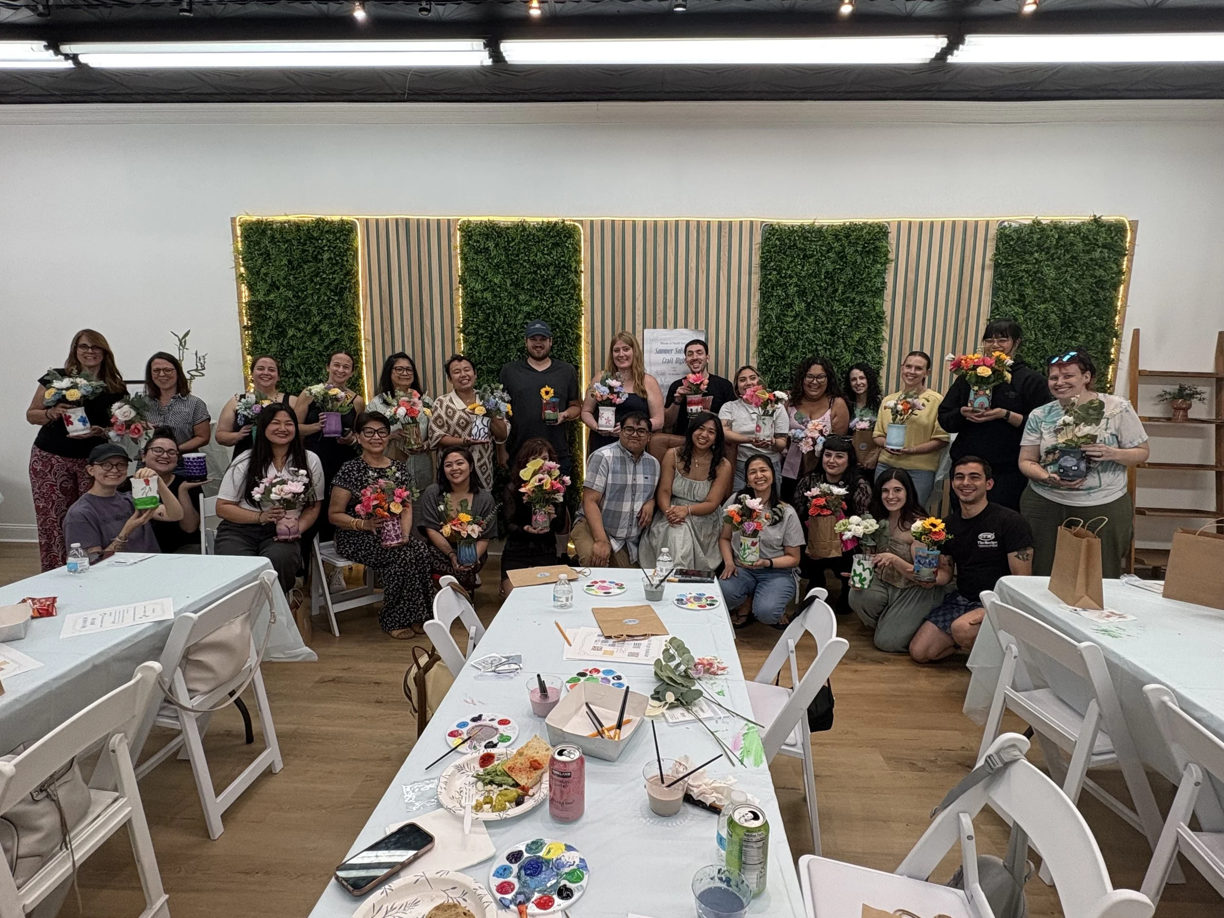 Group of people gathered in a room holding flower arrangements and posing for a photo, with tables set up for art and food, and a decorative green wall in the background.