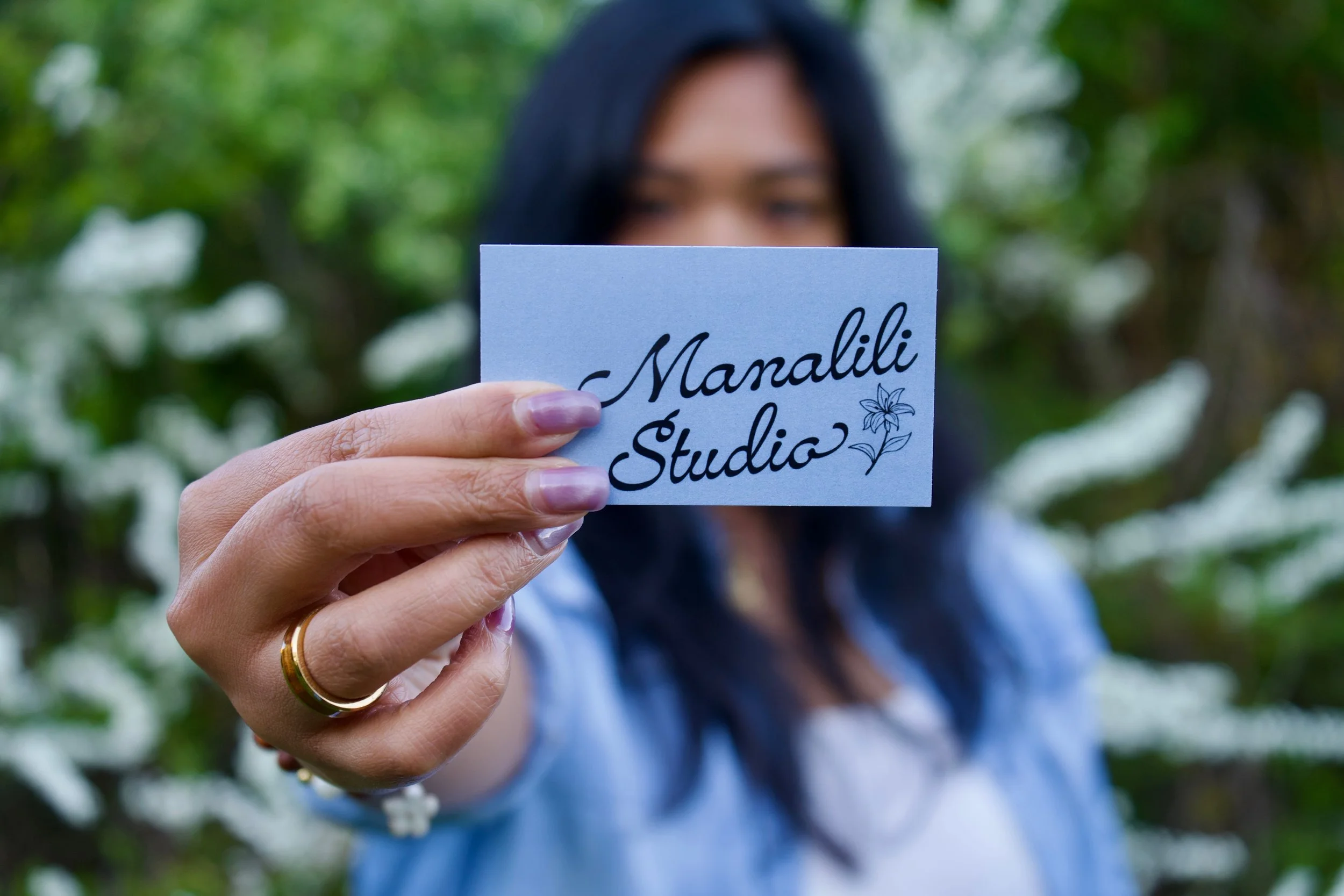 A woman with dark hair holding a business card that reads 'Manalili Studio' with a small flower illustration, outdoors in front of greenery.