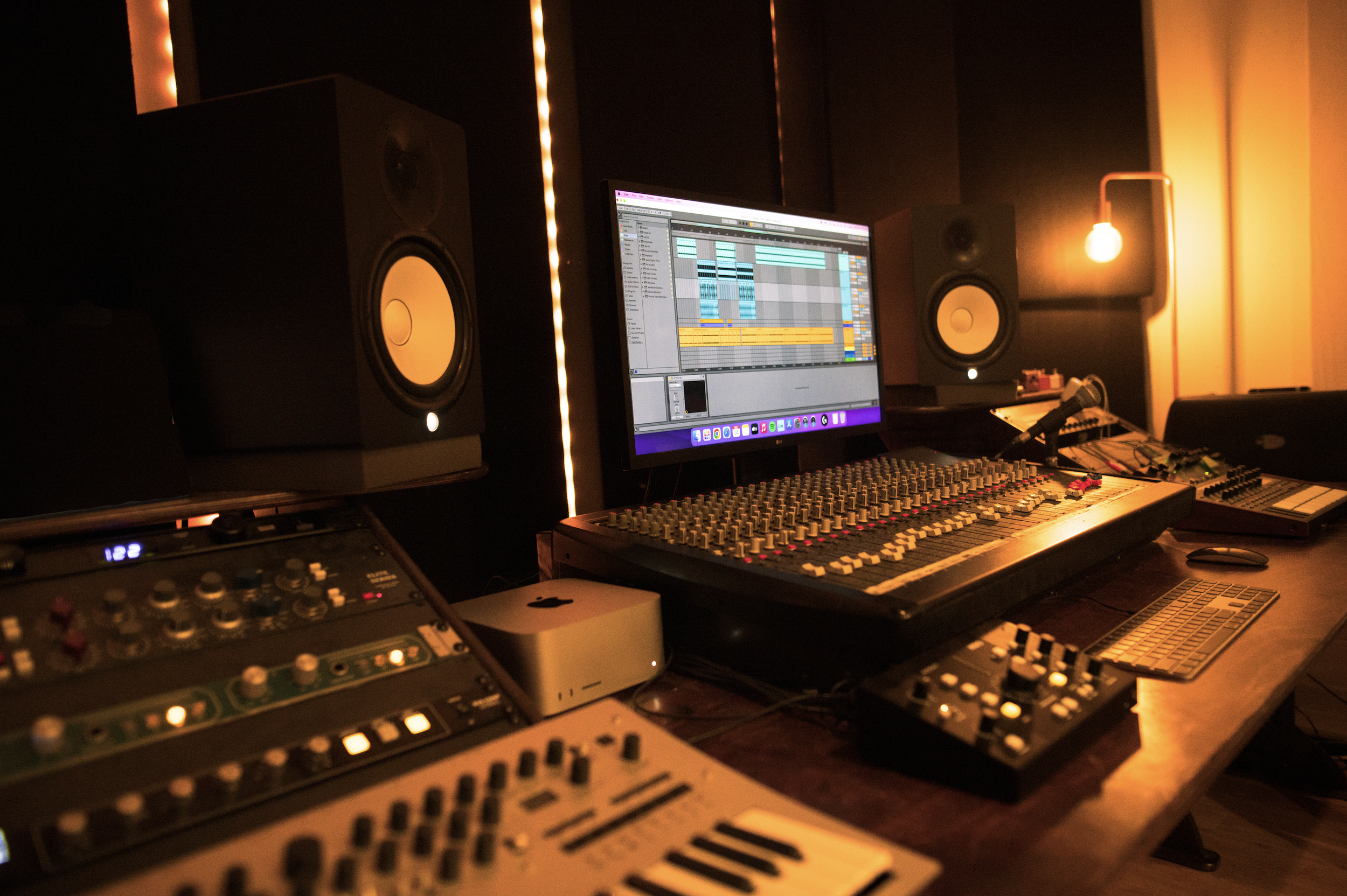 A music production studio with a computer monitor displaying digital audio workstation software, speakers on both sides, a mixing console, and various electronic music equipment illuminated by warm ambient lighting.