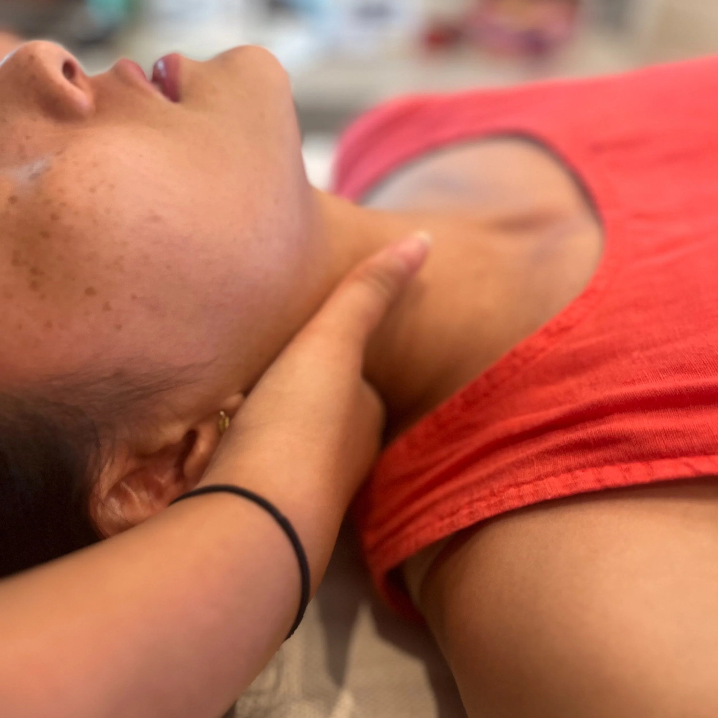 Close-up of a woman lying down, touching her neck with her finger, wearing a red top with a mesh shoulder design.