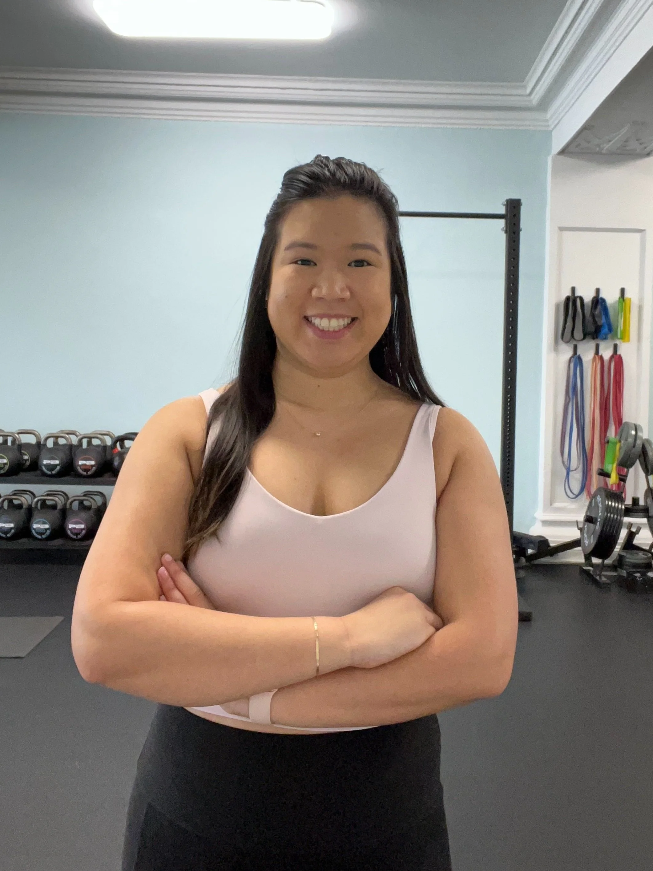 Smiling woman with long dark hair standing arms crossed in a gym, with weights and resistance bands in the background.