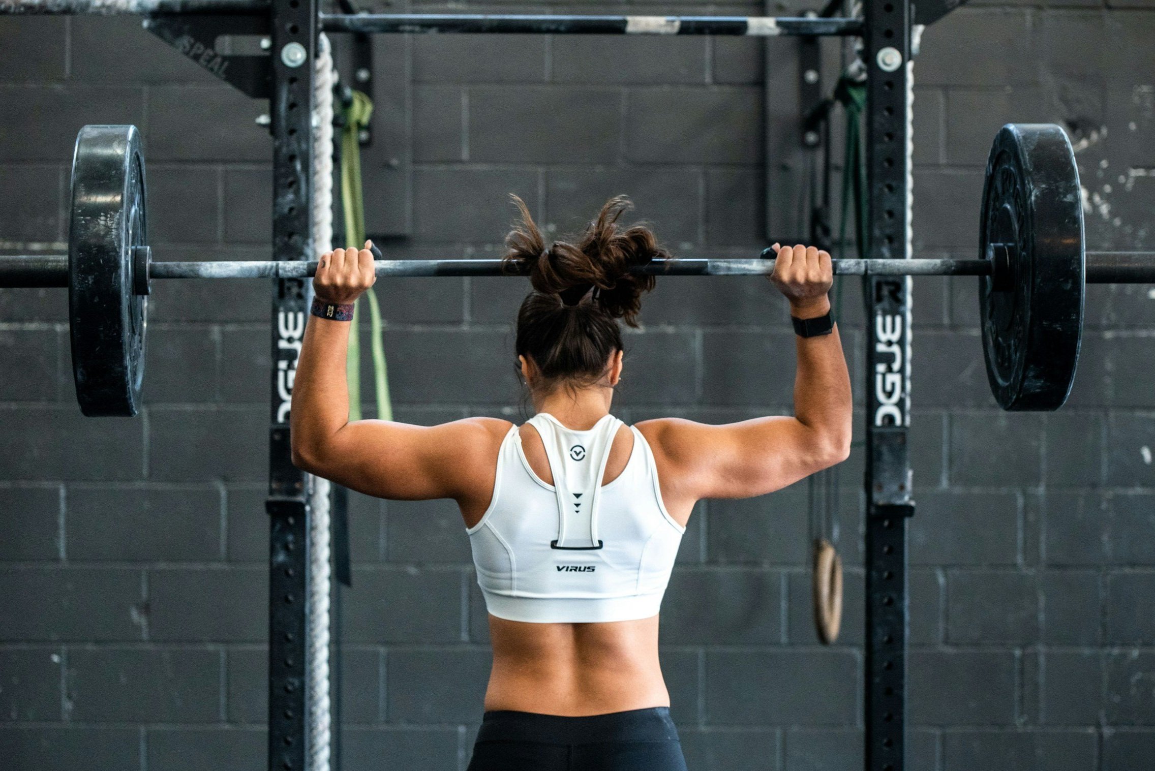 Woman with brown hair in a ponytail lifting a barbell overhead in a gym, wearing a white sports bra and black shorts.