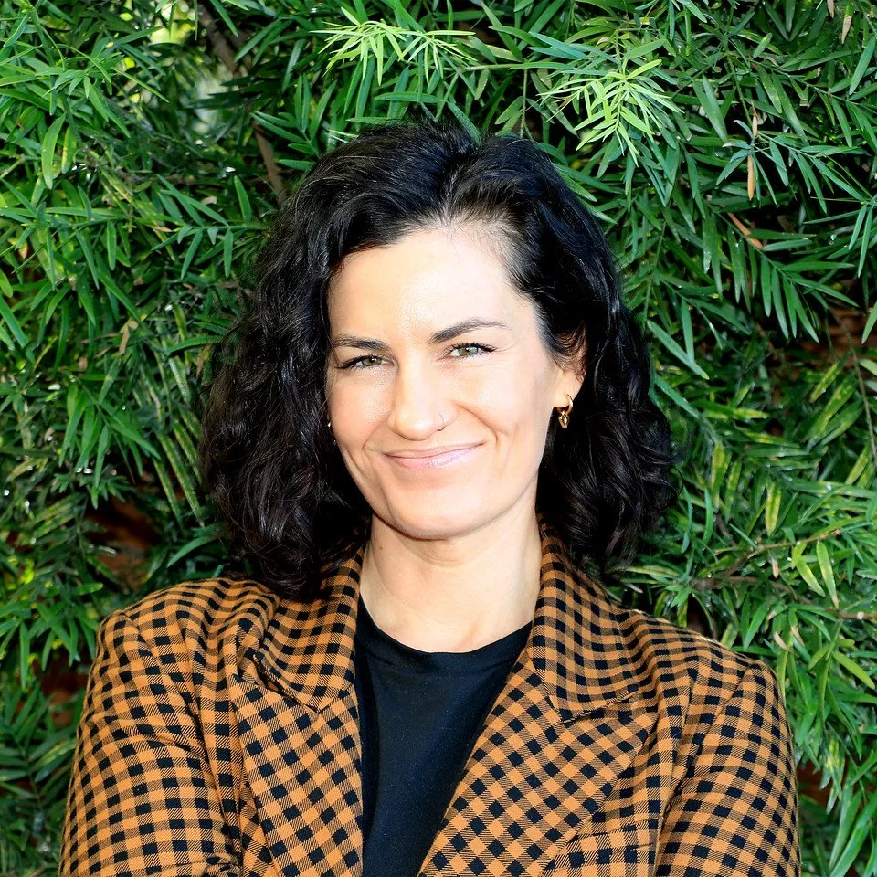 A woman with shoulder-length curly black hair, wearing a brown and black checkered blazer and a black shirt, standing in front of green leafy plants.