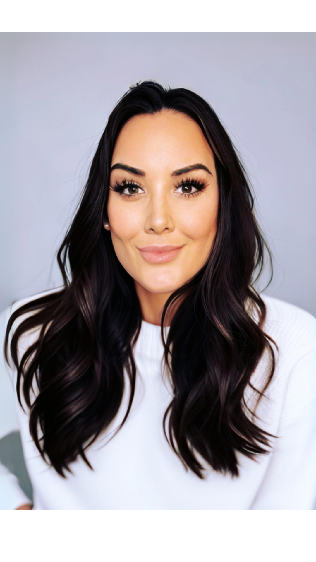 Close-up of a woman with long dark hair, makeup, and a white top, smiling slightly against a plain background.