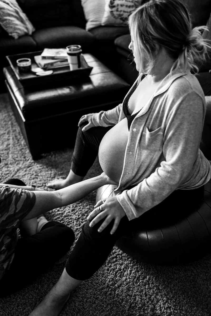 A pregnant mother is sitting on a birth ball during a contraction at her home birth while her midwife is gently feeling the outside of her womb support her in labor.
