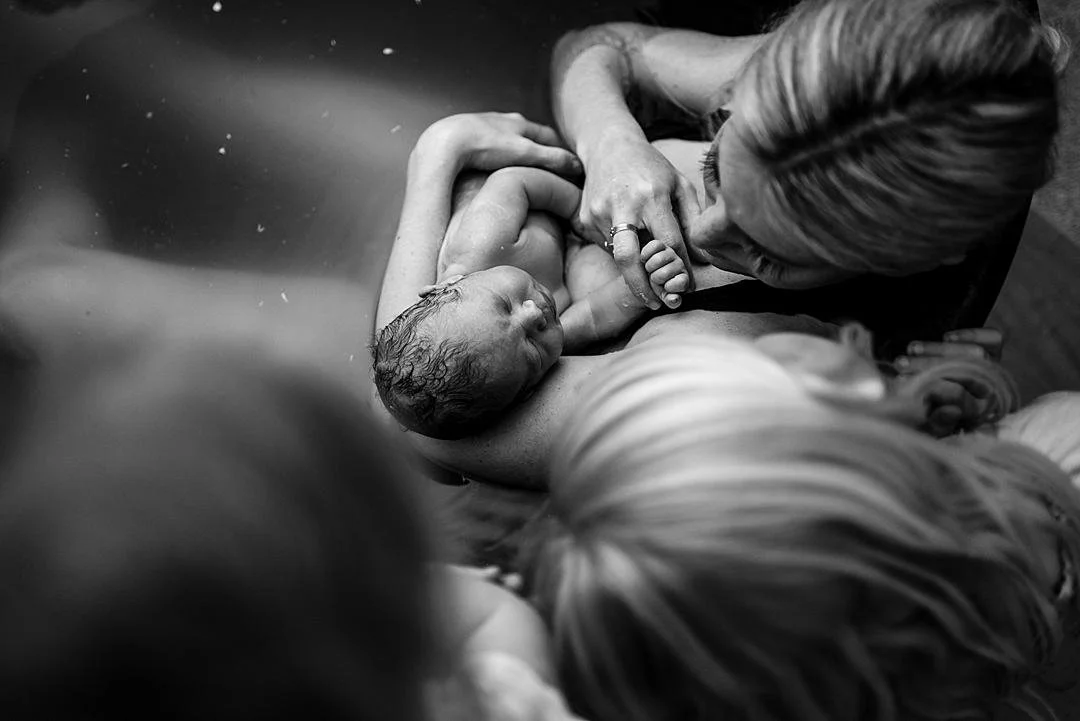 Two young girls peer over the birth tub at the new baby brother who has just been born and is resting in his mothers arms as the enjoy the warmth from the water in the birth pool. The mother gazes at her son after her home water birth and hold his ha