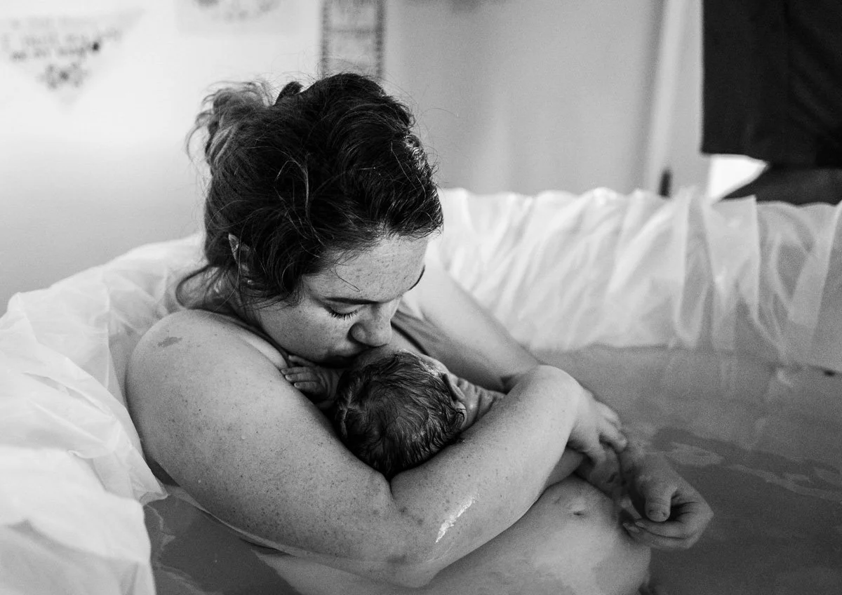 A mother kisses her newborn baby who was just born in a water birth at her homebirth. She is still sitting in the tub with birth affirmations behind her and her soft belly in the water.