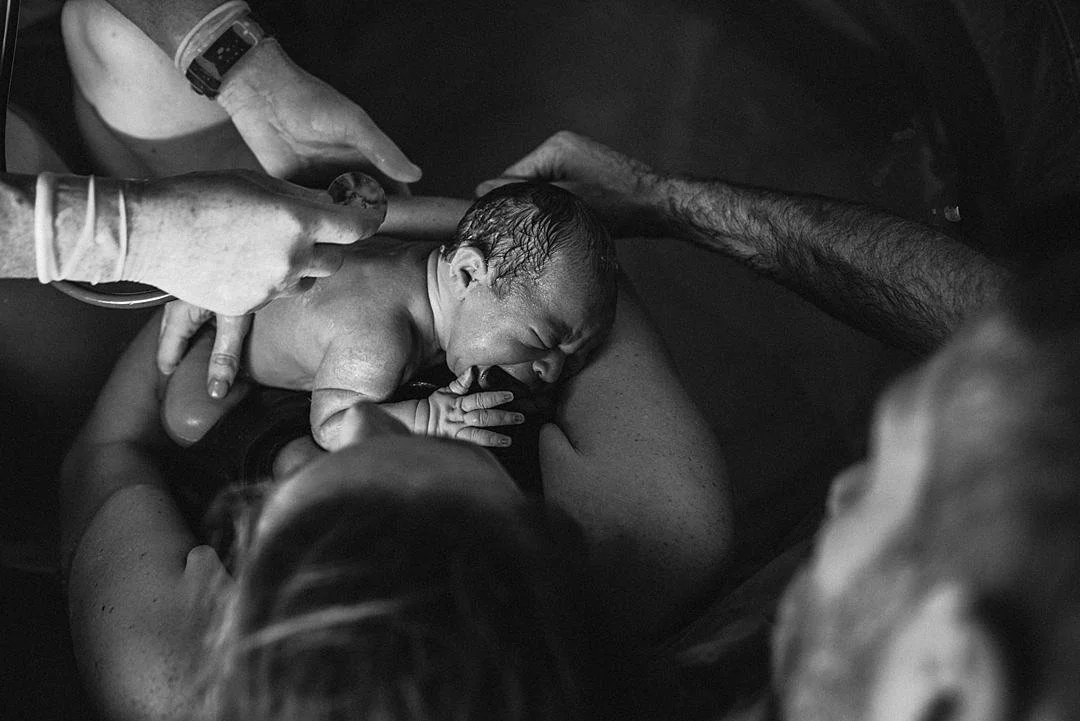 A just born born cries while being held by its mother in a birth pool while the father leans over the pool to watch and puts his arm around the mom and baby while the midwife prepares to listen to the baby with a stethoscope.