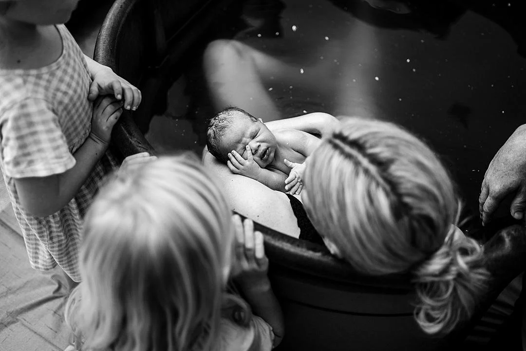Two young sisters peer over the edge of the birth pool to meet their new little brother who is in his mothers arms after just being born. The baby appears to be asleep after his home water birth with his hand laying on his cheek.