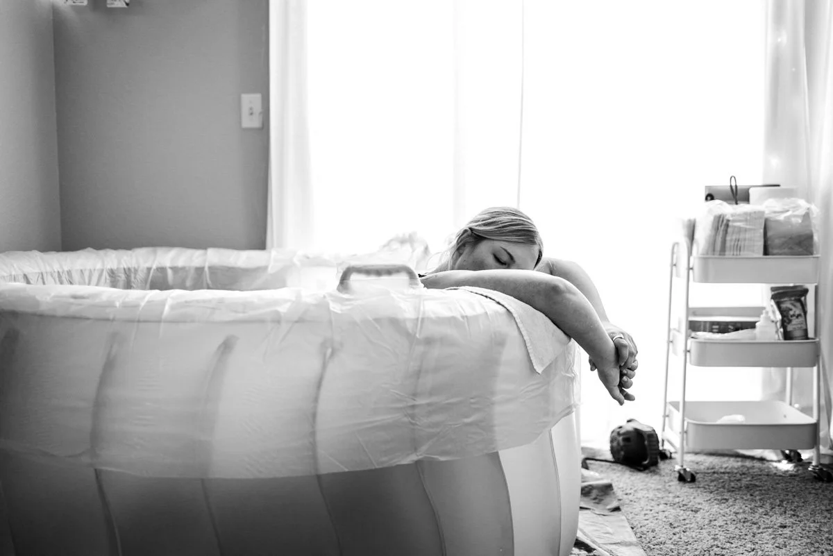 A laboring mom rests on the edge of her birth pool at her home water birth situated in front of a window letting in natural light.