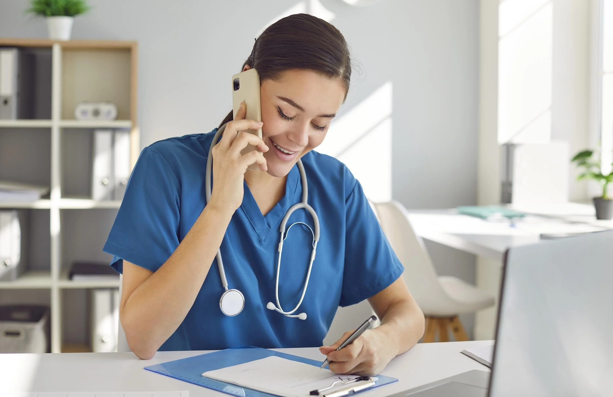 Female healthcare professional in blue scrubs smiling while talking on a smartphone and writing on a clipboard with a pen.