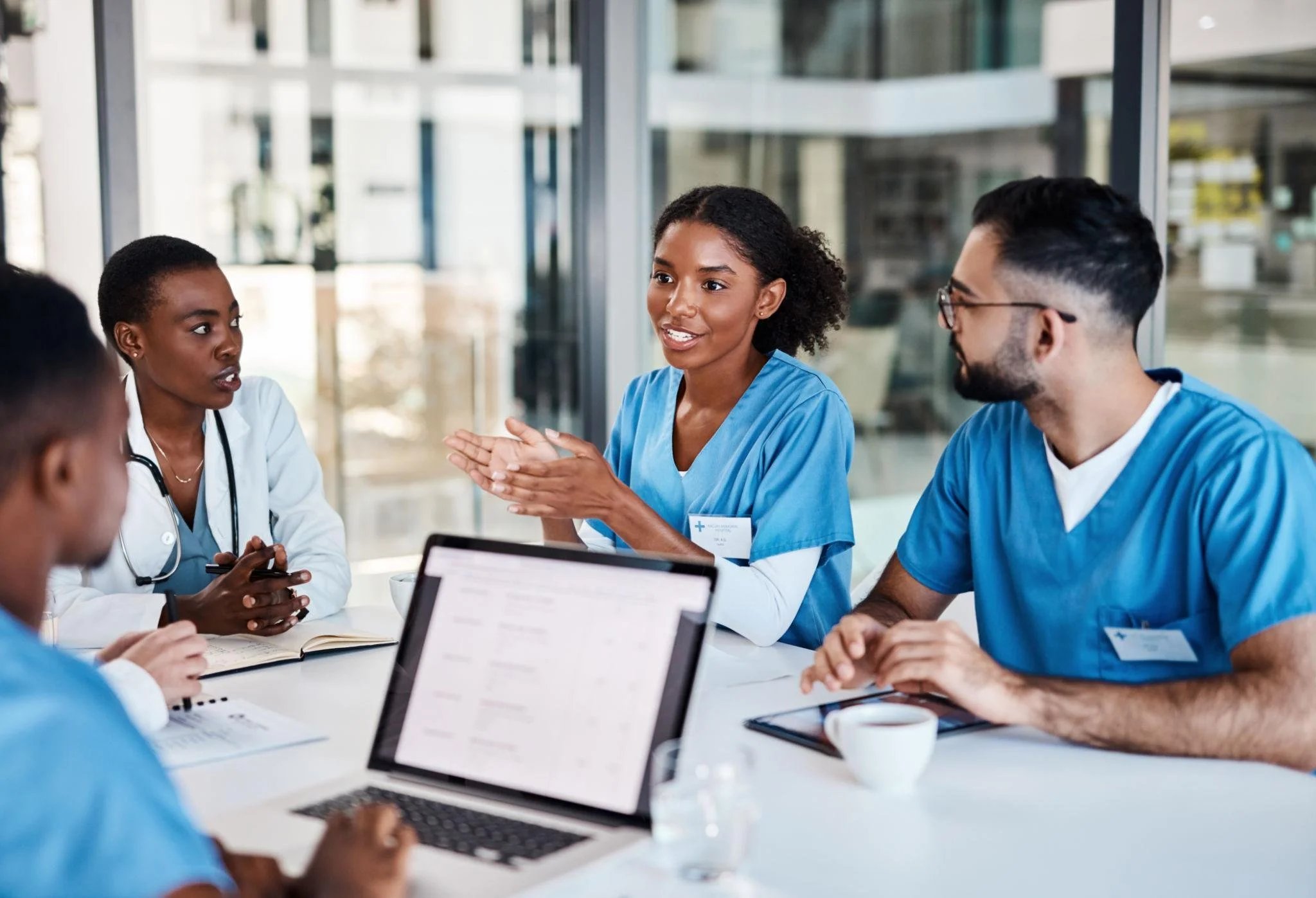 Group of diverse healthcare professionals in discussion at a conference table, with laptops, notebooks, and coffee cups, in a modern office or conference room.
