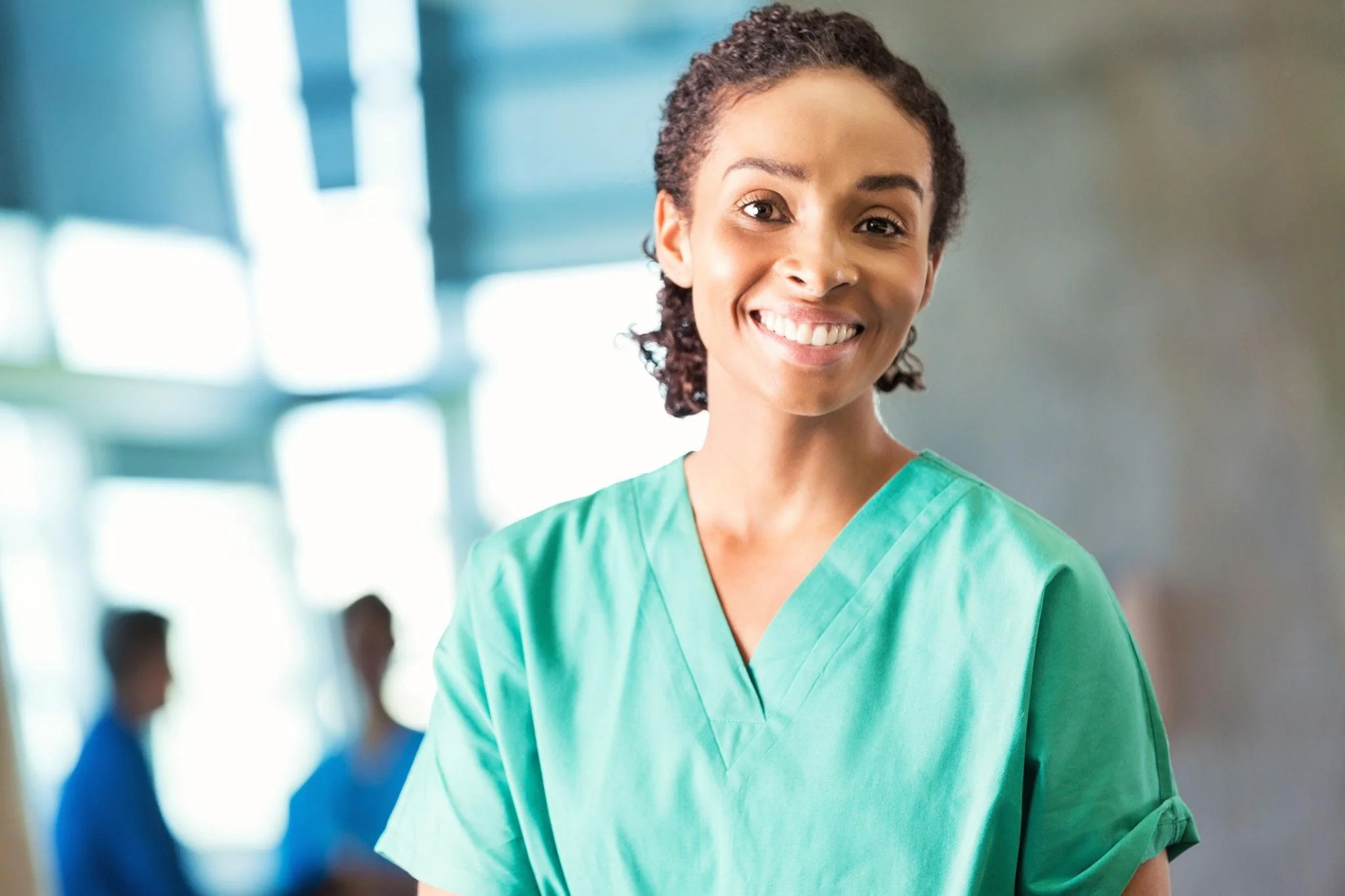 A smiling healthcare worker in green scrubs in a hospital corridor.