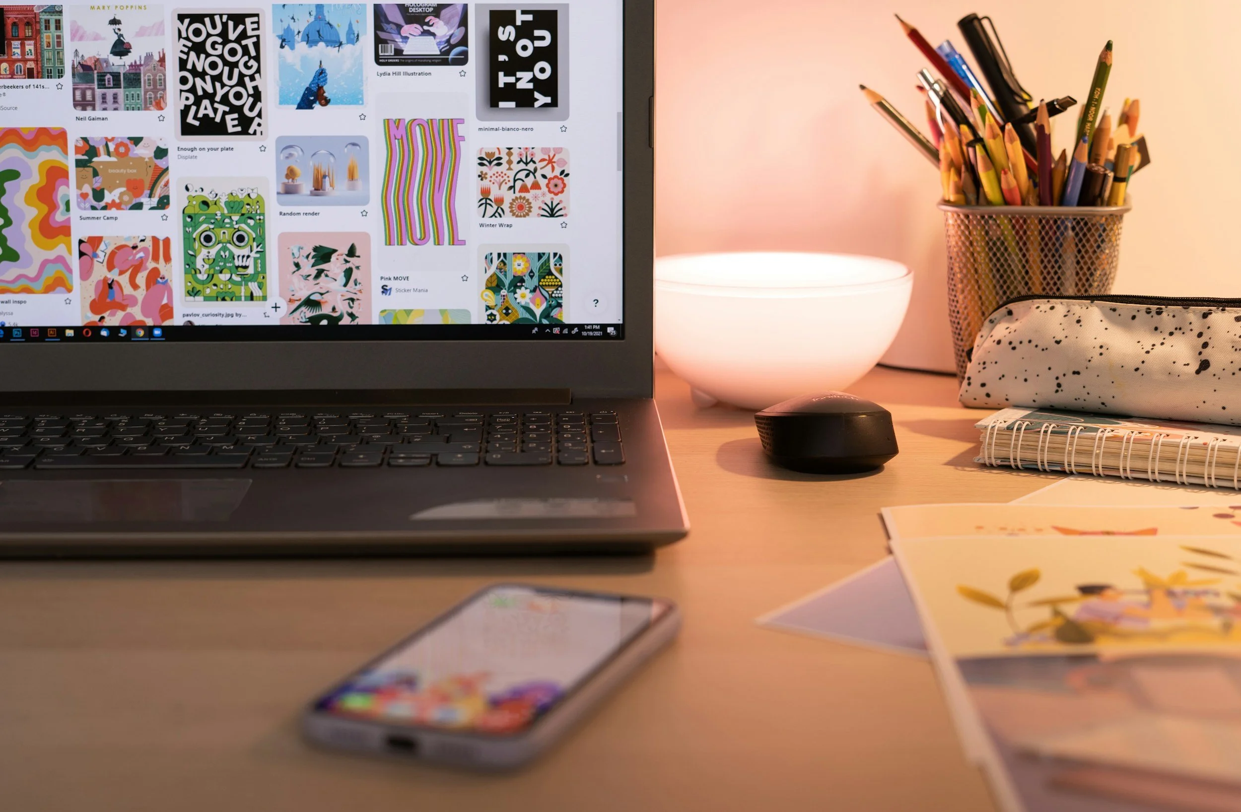 A computer screen displaying colorful digital artwork and illustrations, with a desk that has a small stack of printed artwork, a phone, a keyboard, and a mouse. To the right of the computer, there is a container filled with colored pencils, a lamp, and a speckled pouch. There are also some printed artwork pieces, resembling illustrations or paintings, spread out on the desk.