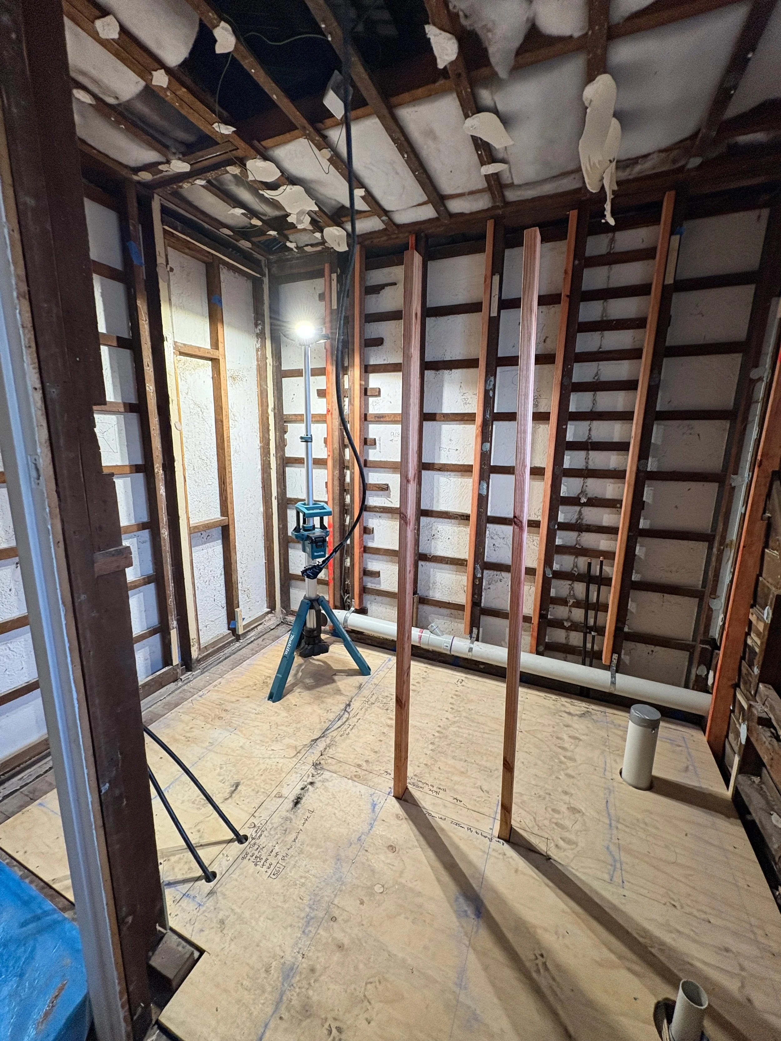Interior of a room under construction, showing exposed wooden framing, plumbing pipes, and a work light.