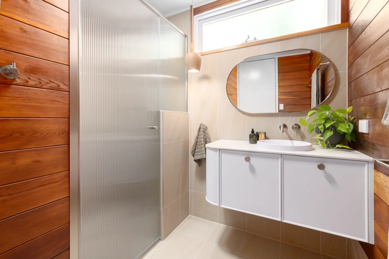 Modern bathroom with wooden wall paneling, a frosted glass shower door, a white vanity with a potted plant, an oval mirror, and a window.