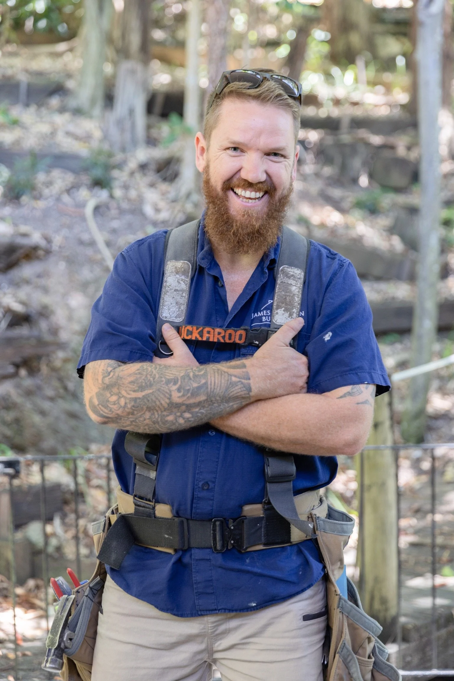 A smiling man with a beard and tattoos on his arm, dressed in a blue shirt, with a backpack, standing outdoors in a wooded area.