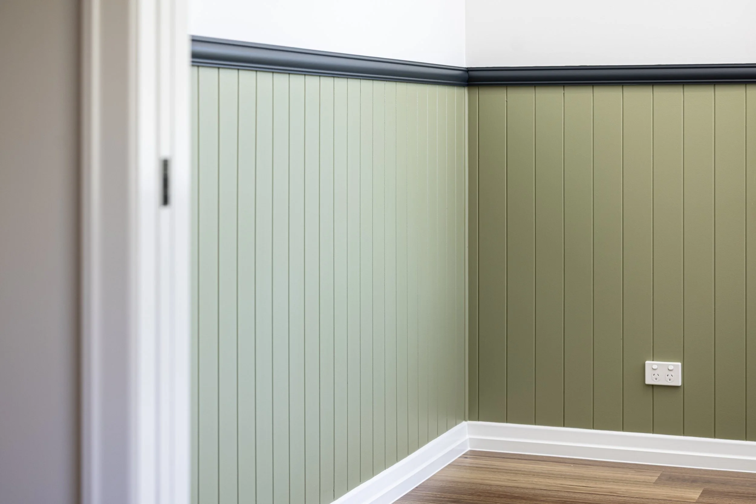 Interior corner of a room with olive green vertical paneling walls, white baseboard, a black electrical outlet, and laminate wooden flooring.