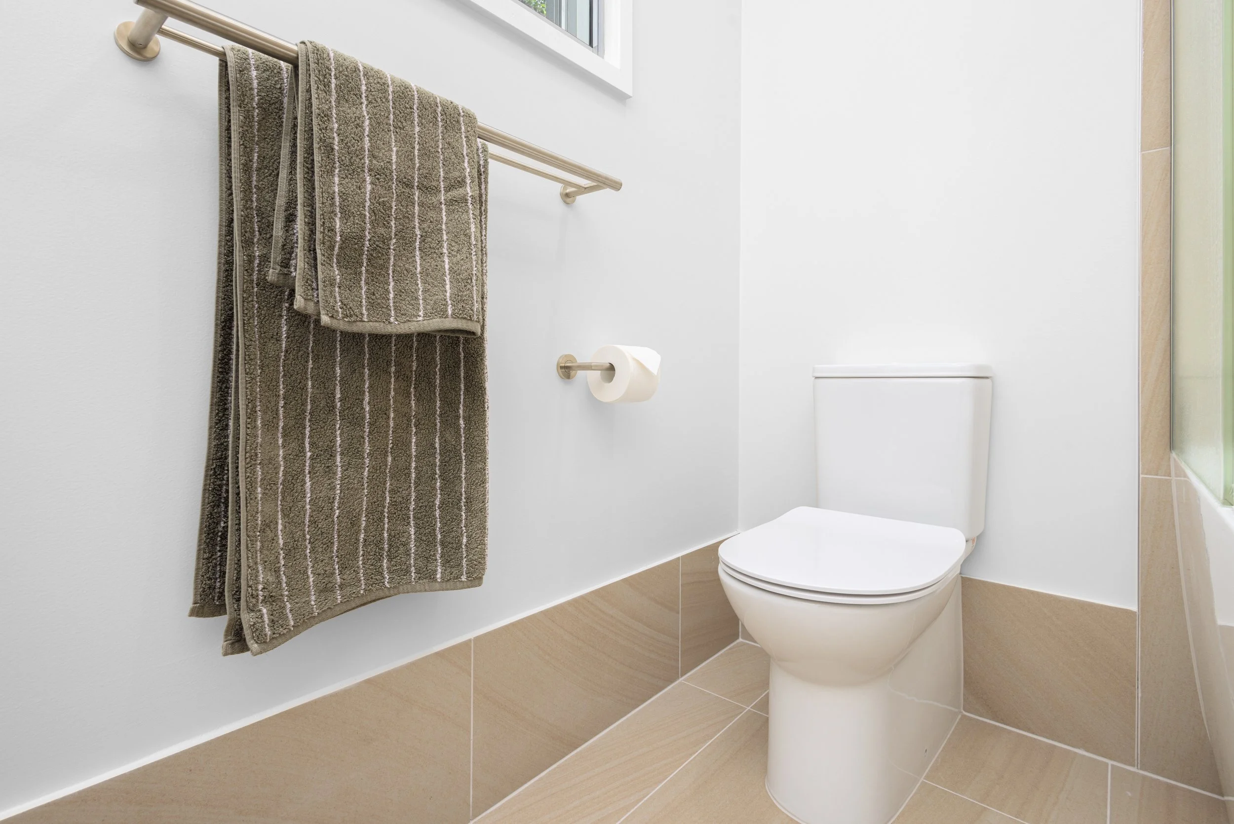 A clean bathroom with a white toilet, brown striped towels hanging on a metal towel rack, and a window above the towels.